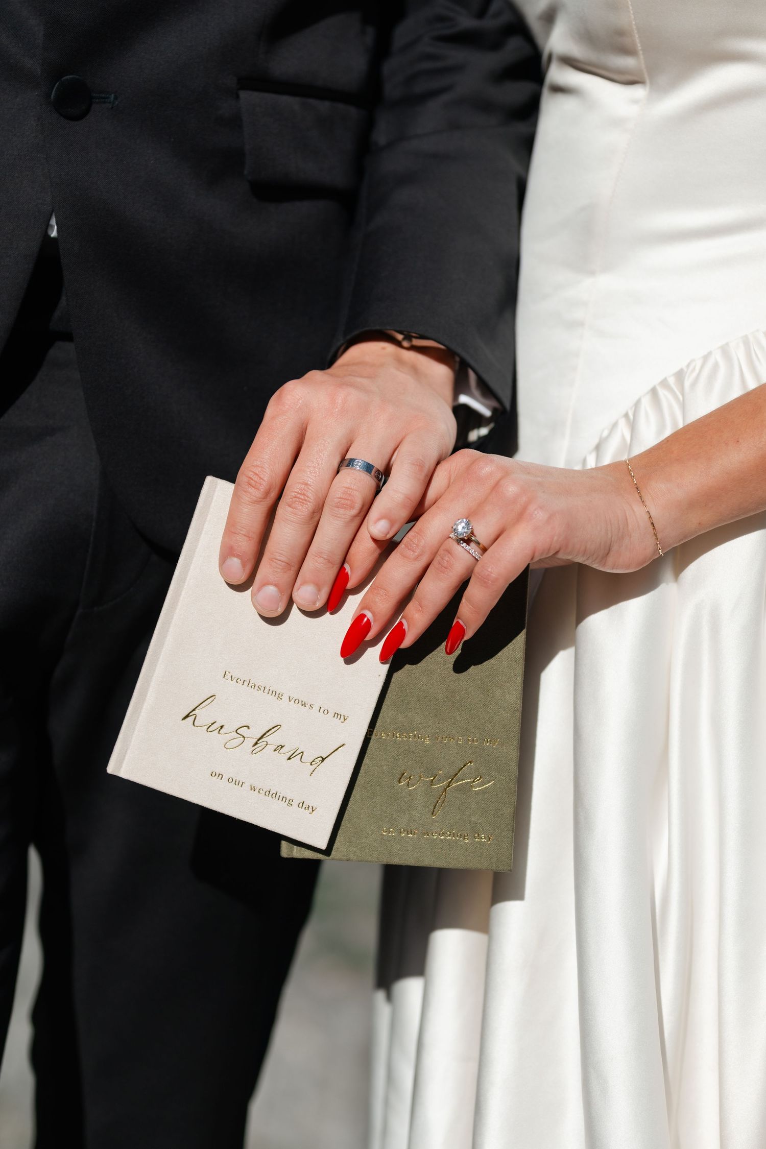 Newlyweds holding wedding vow books, bride with red nails and diamond ring, groom in black suit with gold band.