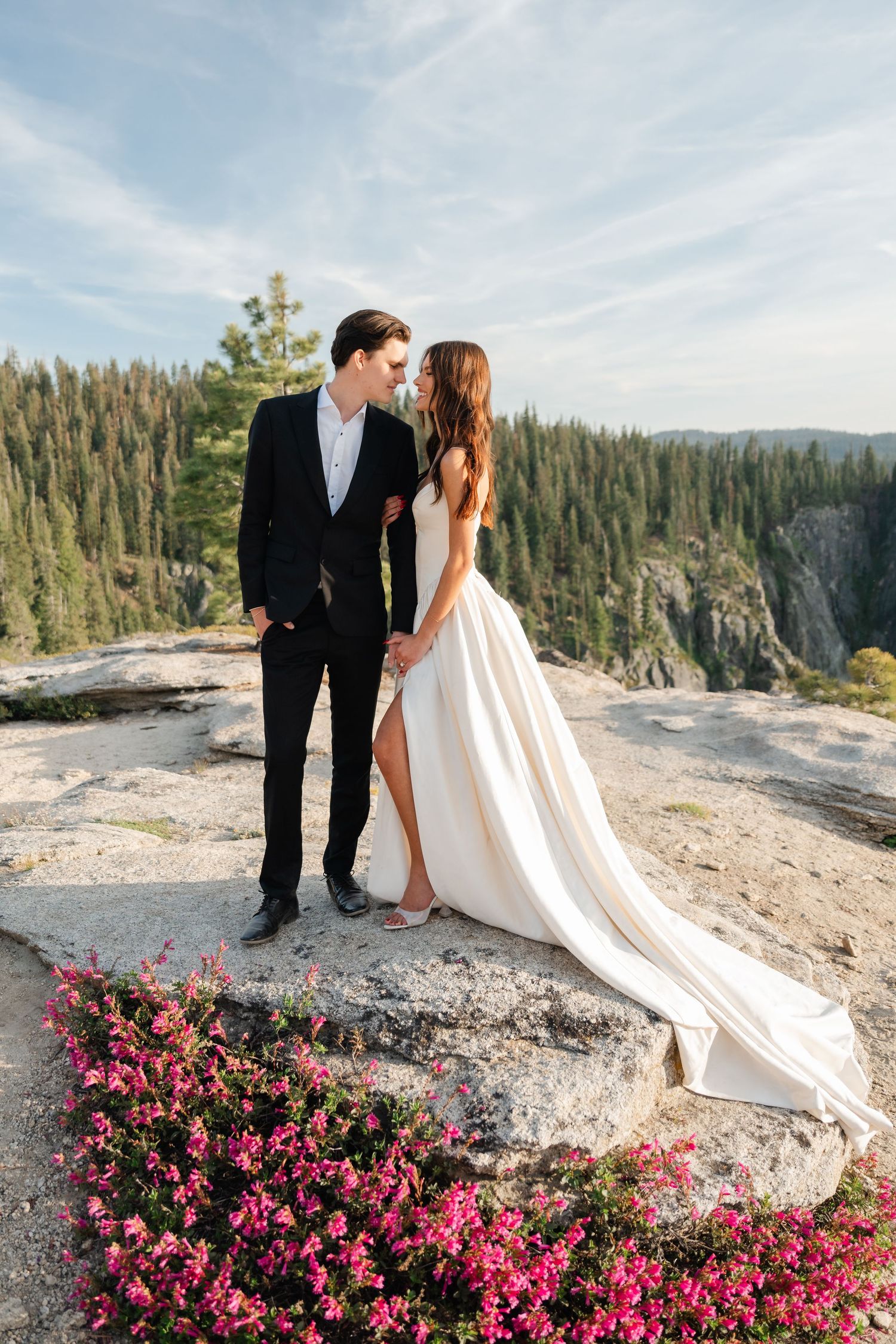 Bride and groom share a romantic moment on a rocky mountain overlook with pink wildflowers and pine forest backdrop.