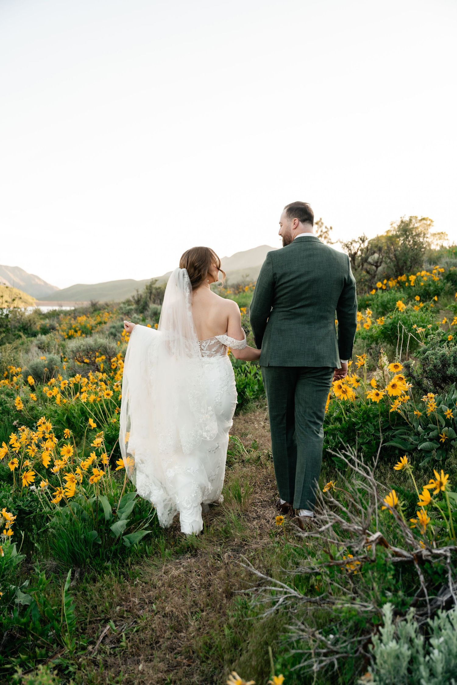 Newlywed couple walks through field of yellow wildflowers with mountain lake backdrop.