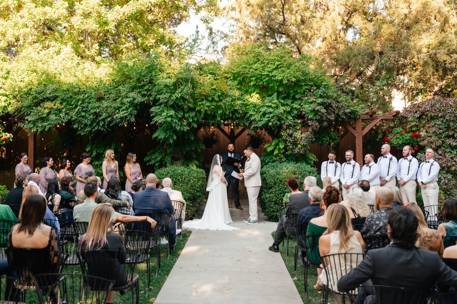 Outdoor garden wedding ceremony with guests seated on black chairs and greenery-covered arch.