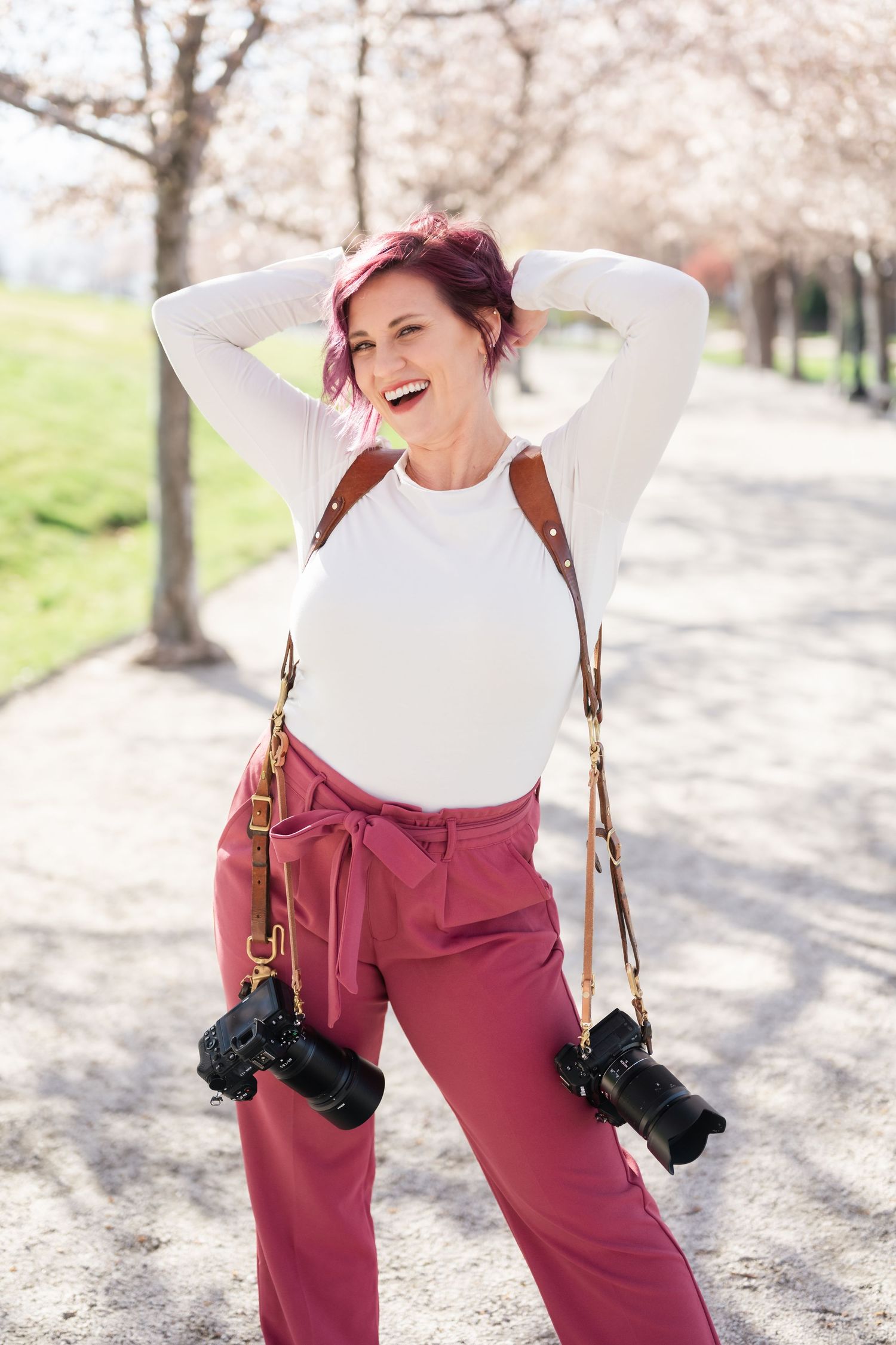 Photographer in pink pants and white top stands joyfully under cherry blossoms with cameras hanging from shoulders.
