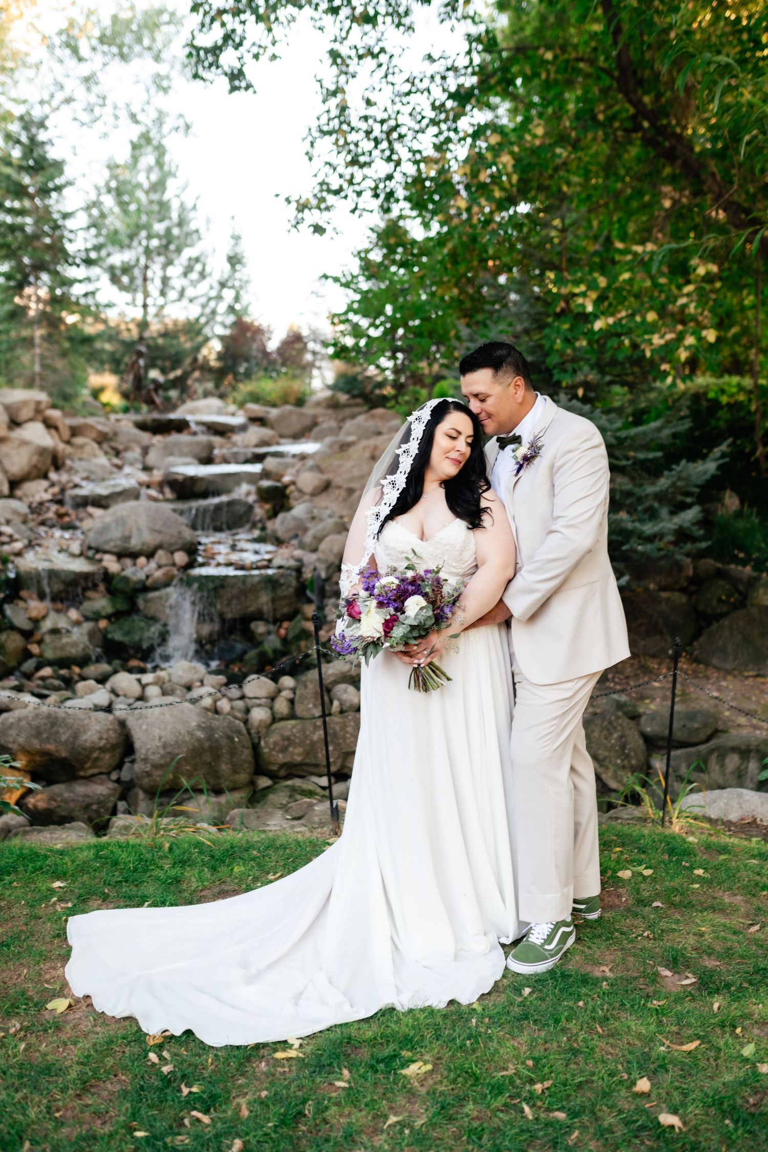 A couple poses together by a cascading rock waterfall garden during their wedding day, with purple florals in the bouquet.