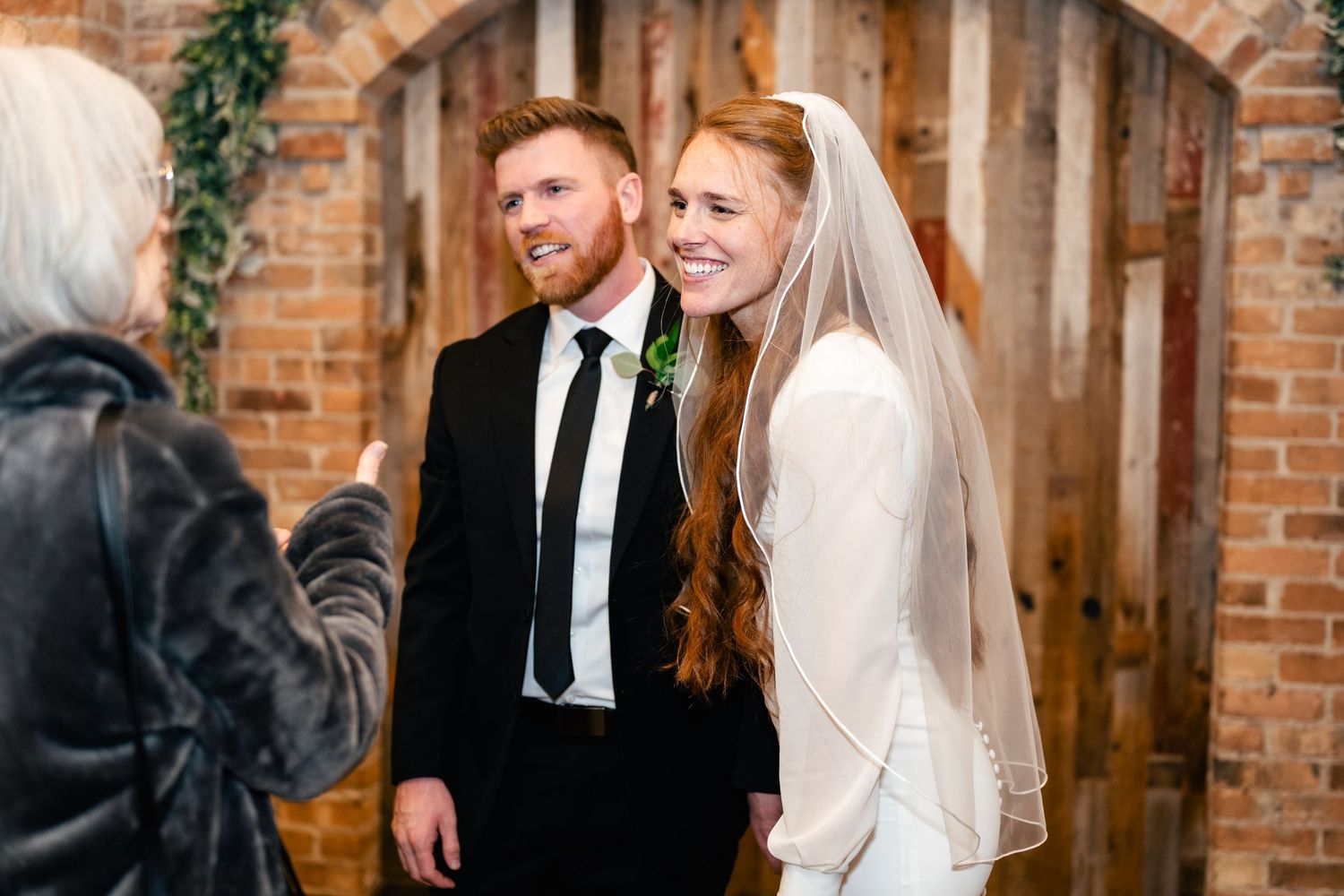 A newly married couple shares a joyful moment in front of a rustic brick wall with wooden accents.