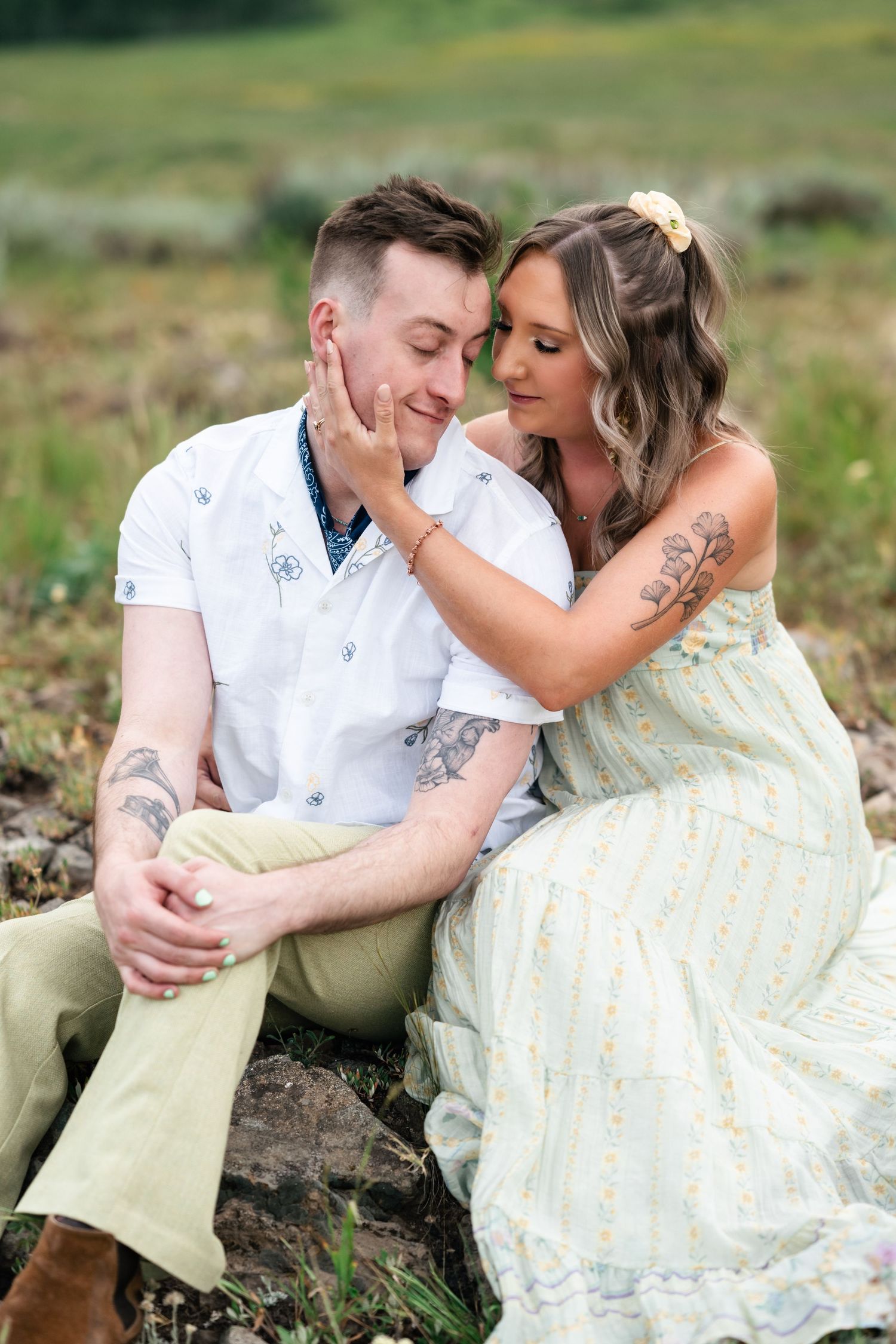 A couple in light summer attire share an intimate moment while sitting together outdoors in a grassy field. Park City engagement photos