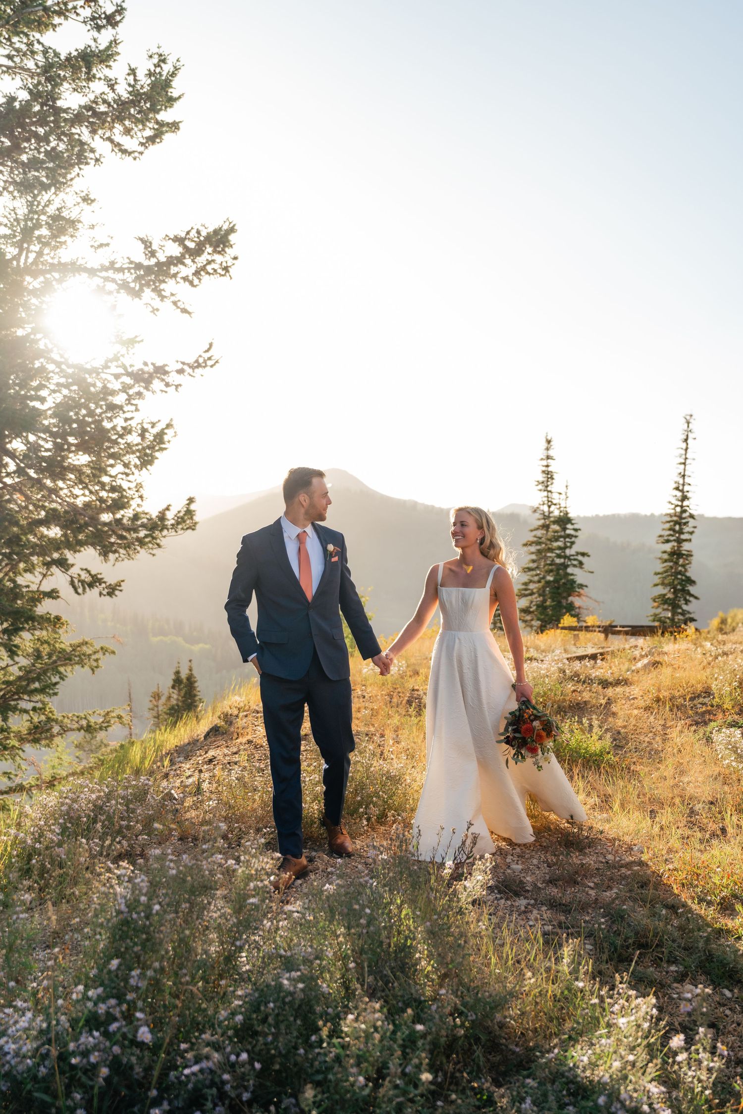 A couple walking hand in hand on a mountain trail at sunset with evergreen trees and golden light in the background. Park City Wedding Photo