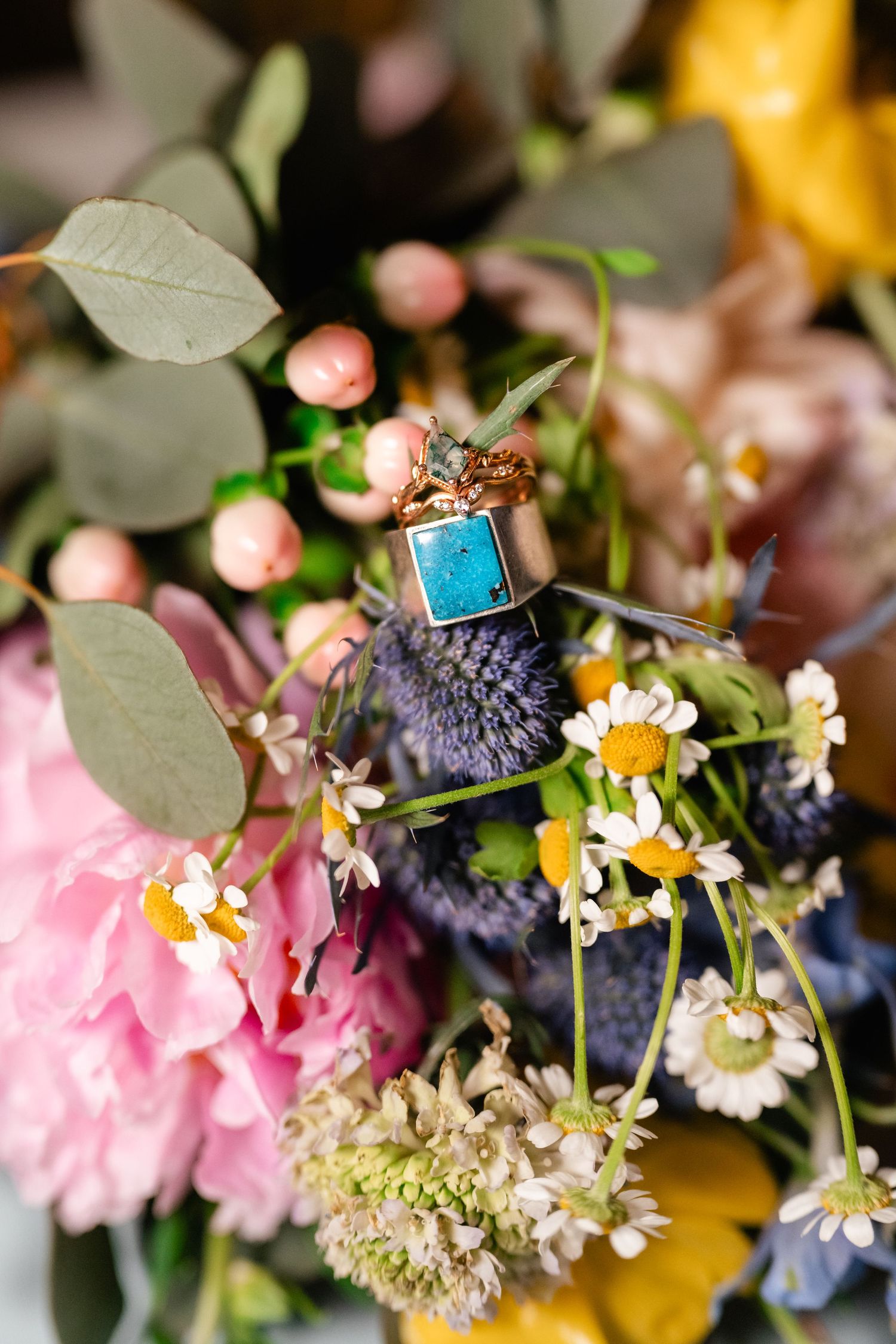 Wedding rings with turquoise stone detail resting on colorful bouquet with peonies, daisies and eucalyptus leaves.