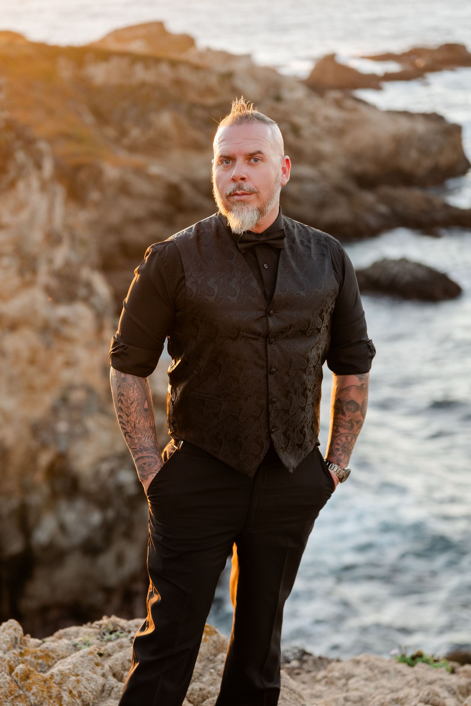 Person in formal black attire poses on coastal rocks at sunset.