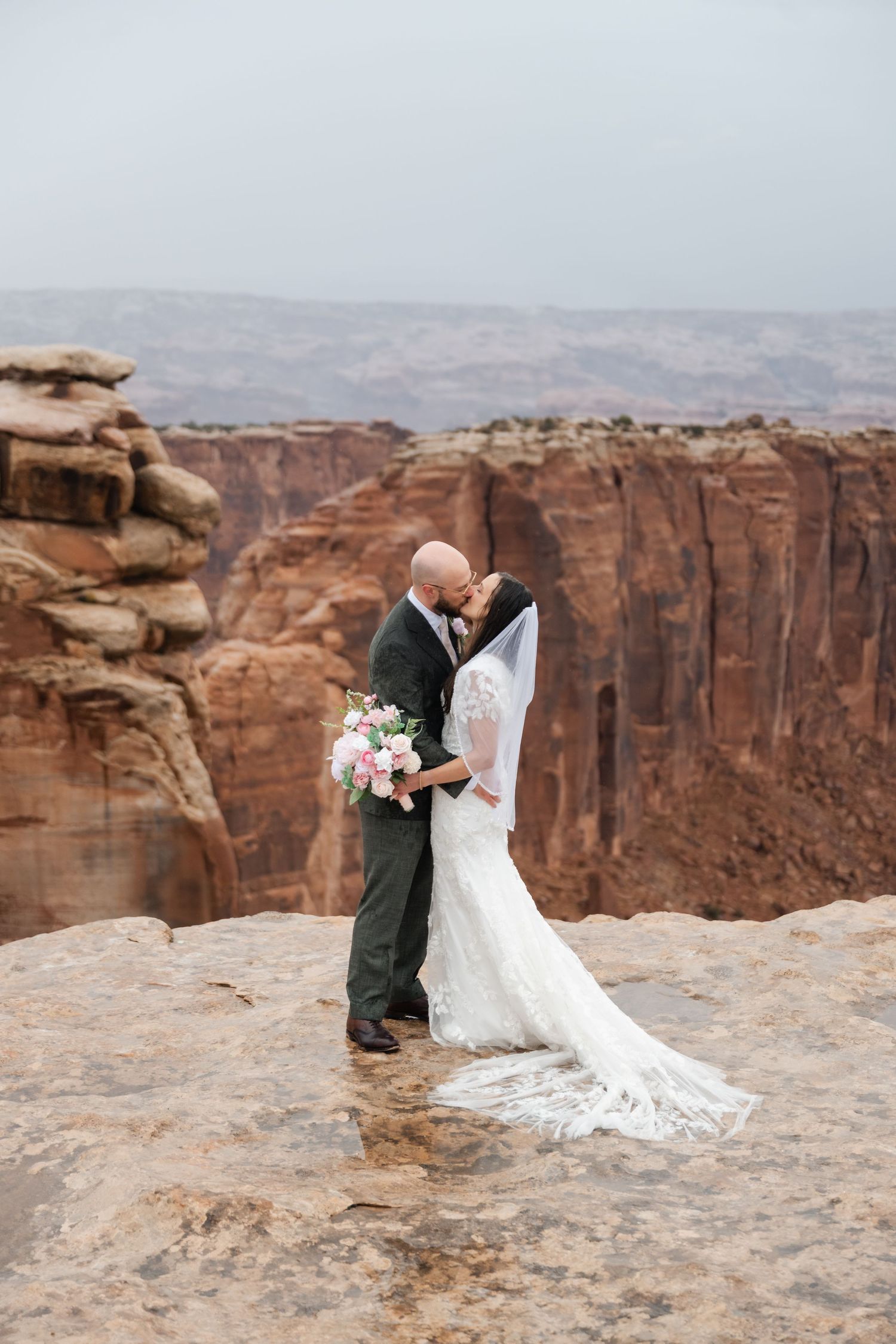 A wedding couple embraces on the edge of majestic red rock cliffs at sunset.