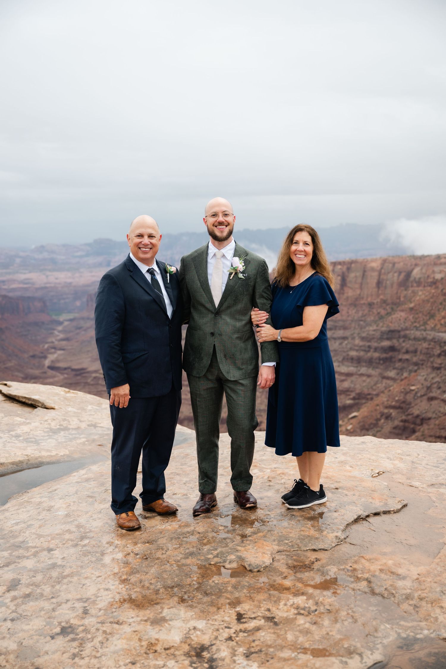 Three people stand together at a scenic overlook with red rock canyon cliffs and desert landscape in the background.