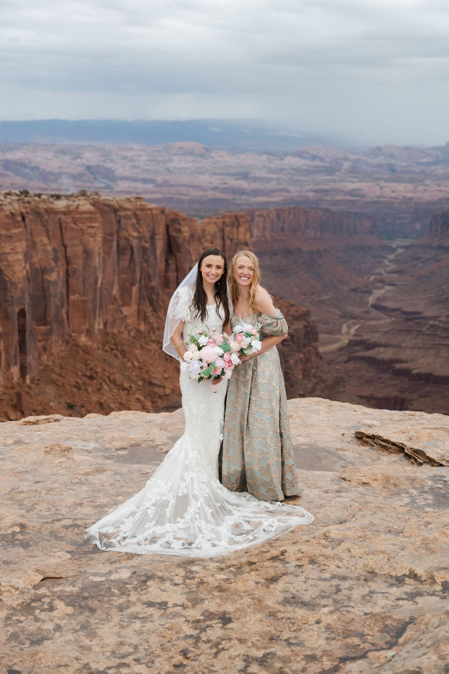 Two bridesmaids in sparkly dresses pose with bouquets on canyon overlook with vast desert views.