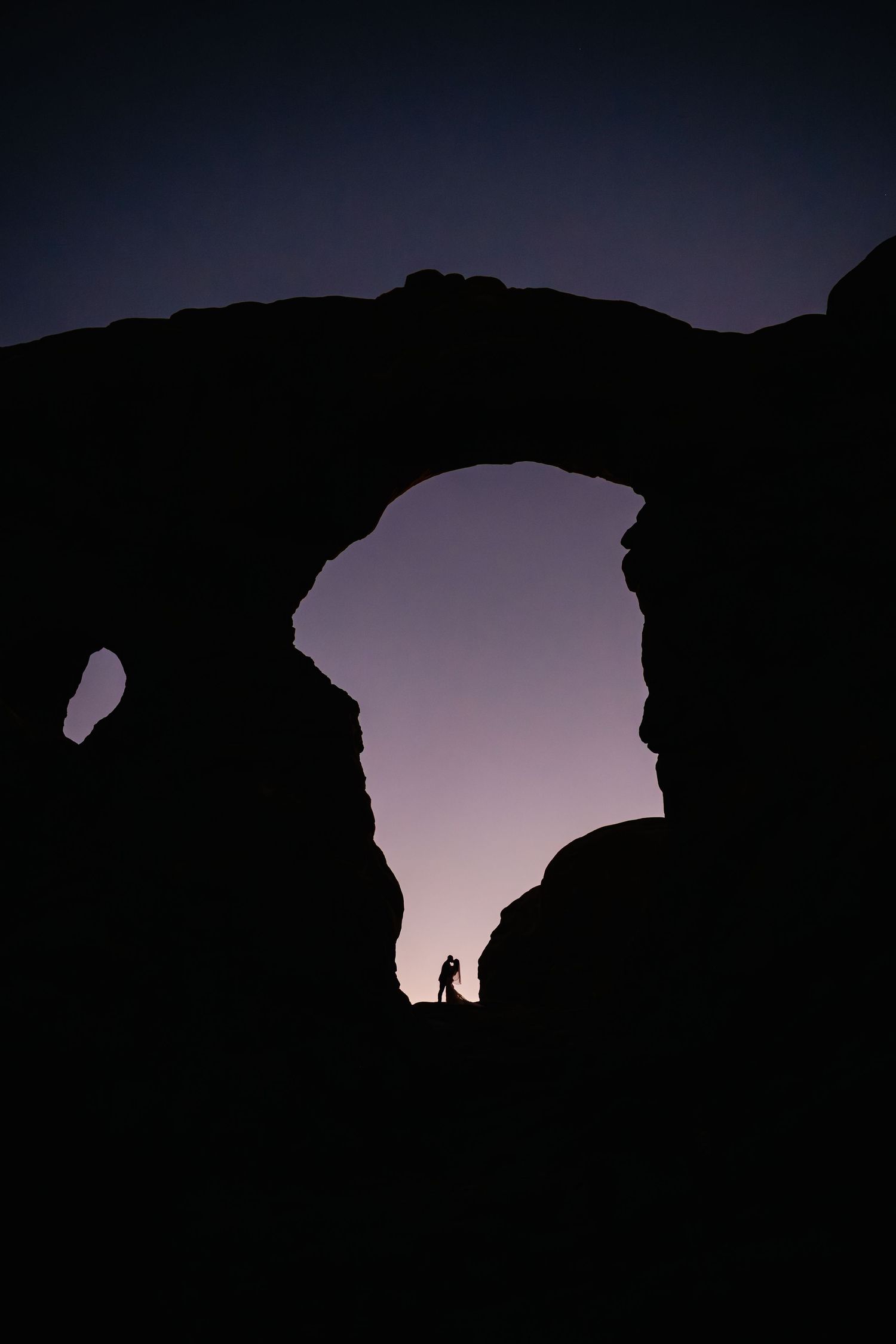 Natural stone arches silhouetted against a dusky purple sky in a desert setting.