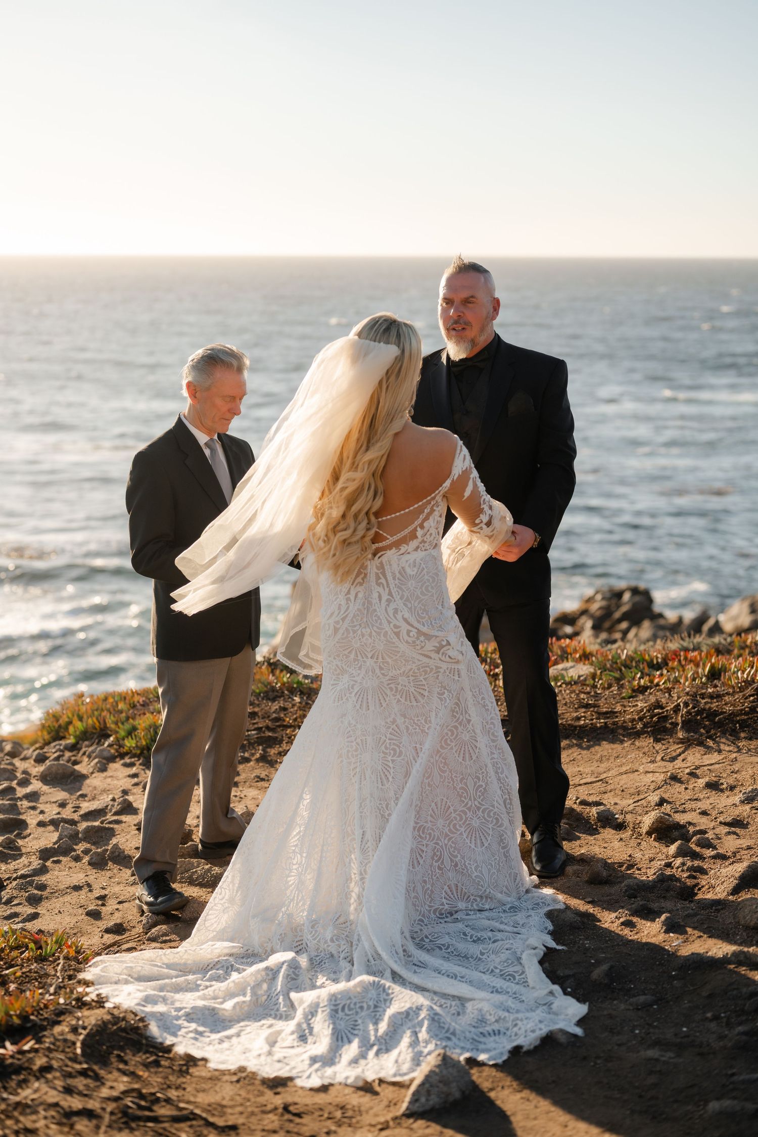 A romantic wedding ceremony takes place on a scenic coastal cliff at sunset with the ocean in the background.