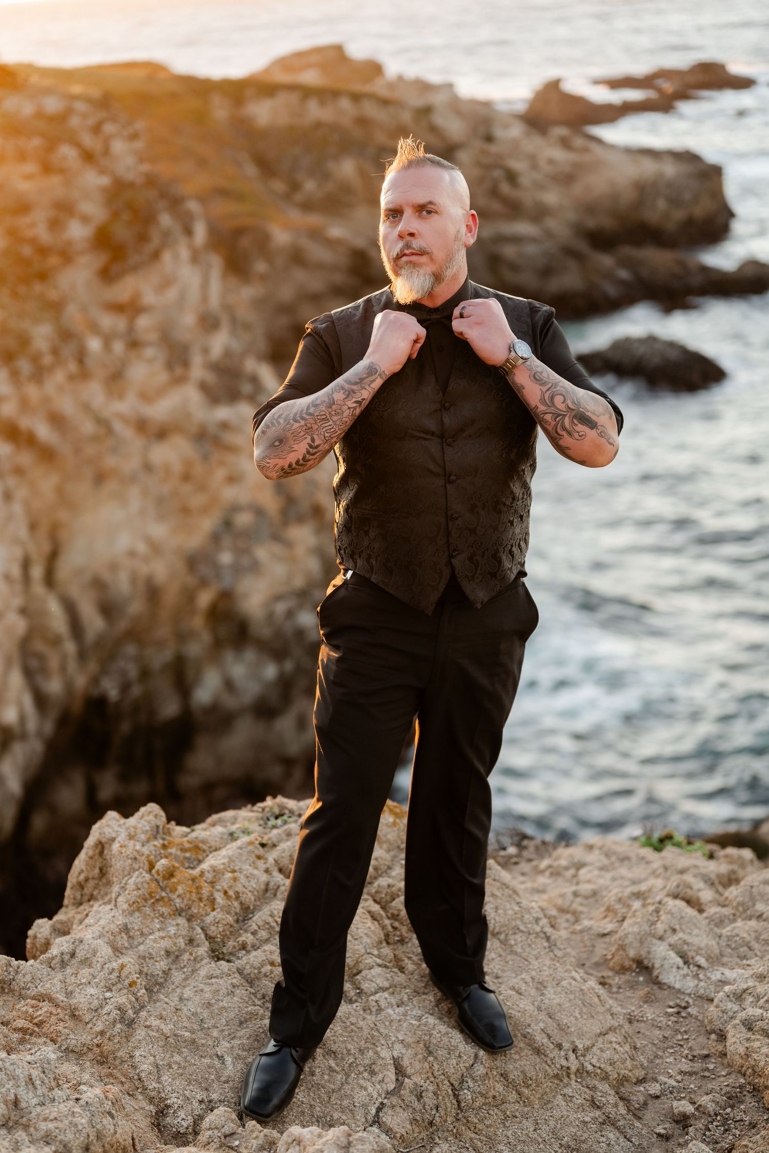 A formal wedding portrait session on rocky coastal cliffs overlooking the ocean at sunset.