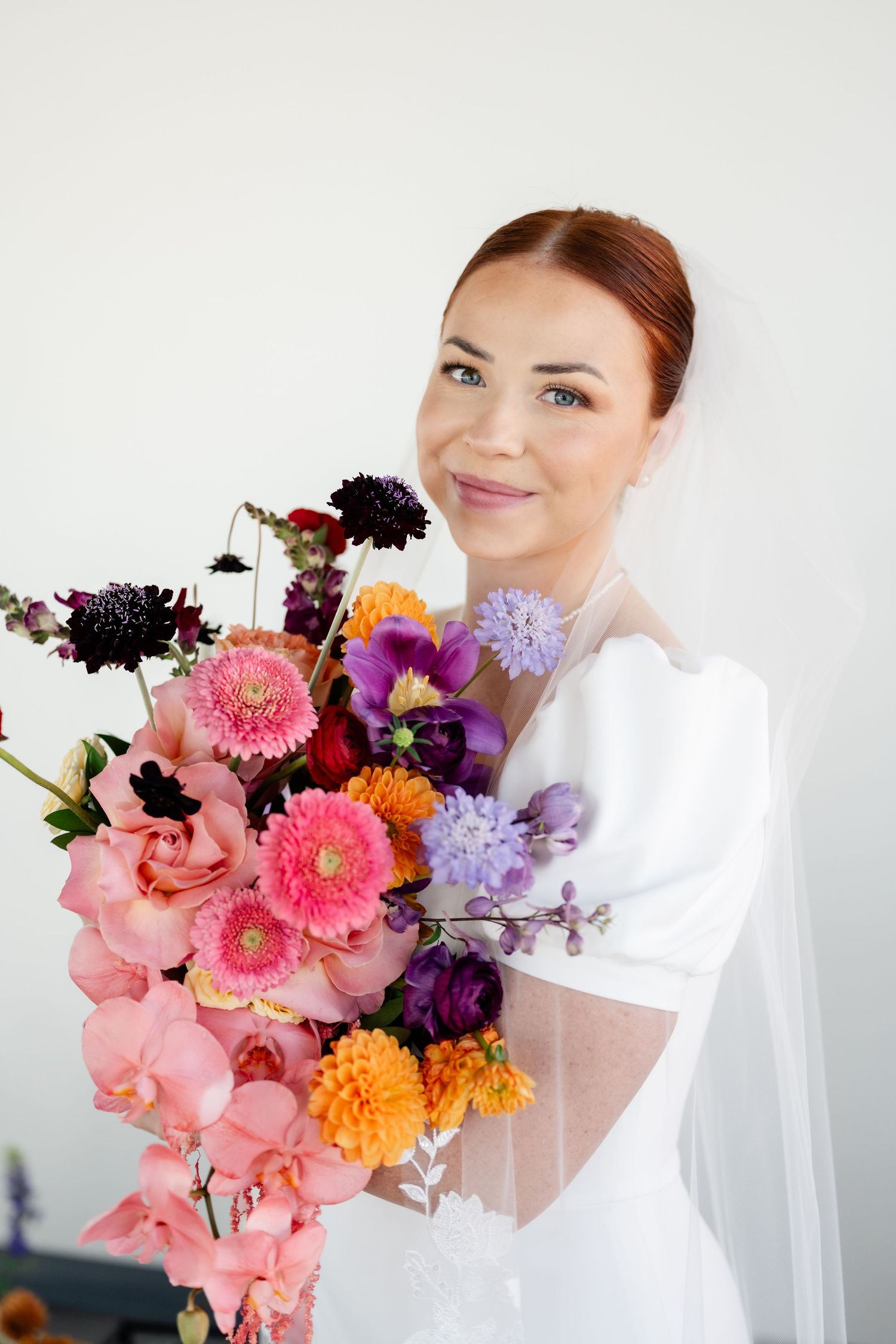 A beautiful bridal bouquet featuring pink orchids, gerbera daisies, purple dahlias and orange marigolds against a white background.