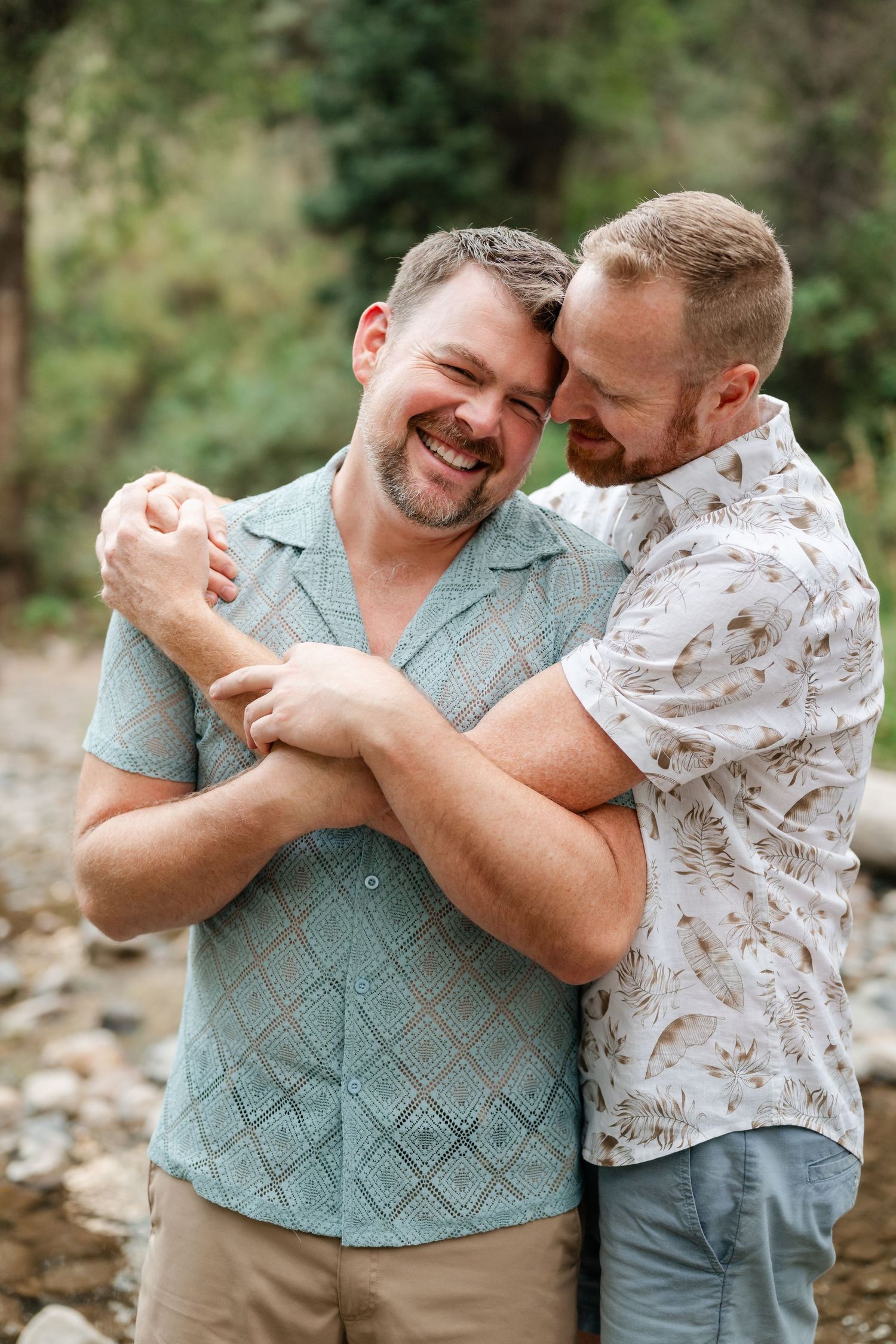 Gay couple engagement photos. Two men embracing and smiling outdoors near rocky terrain with lush green trees in the background.