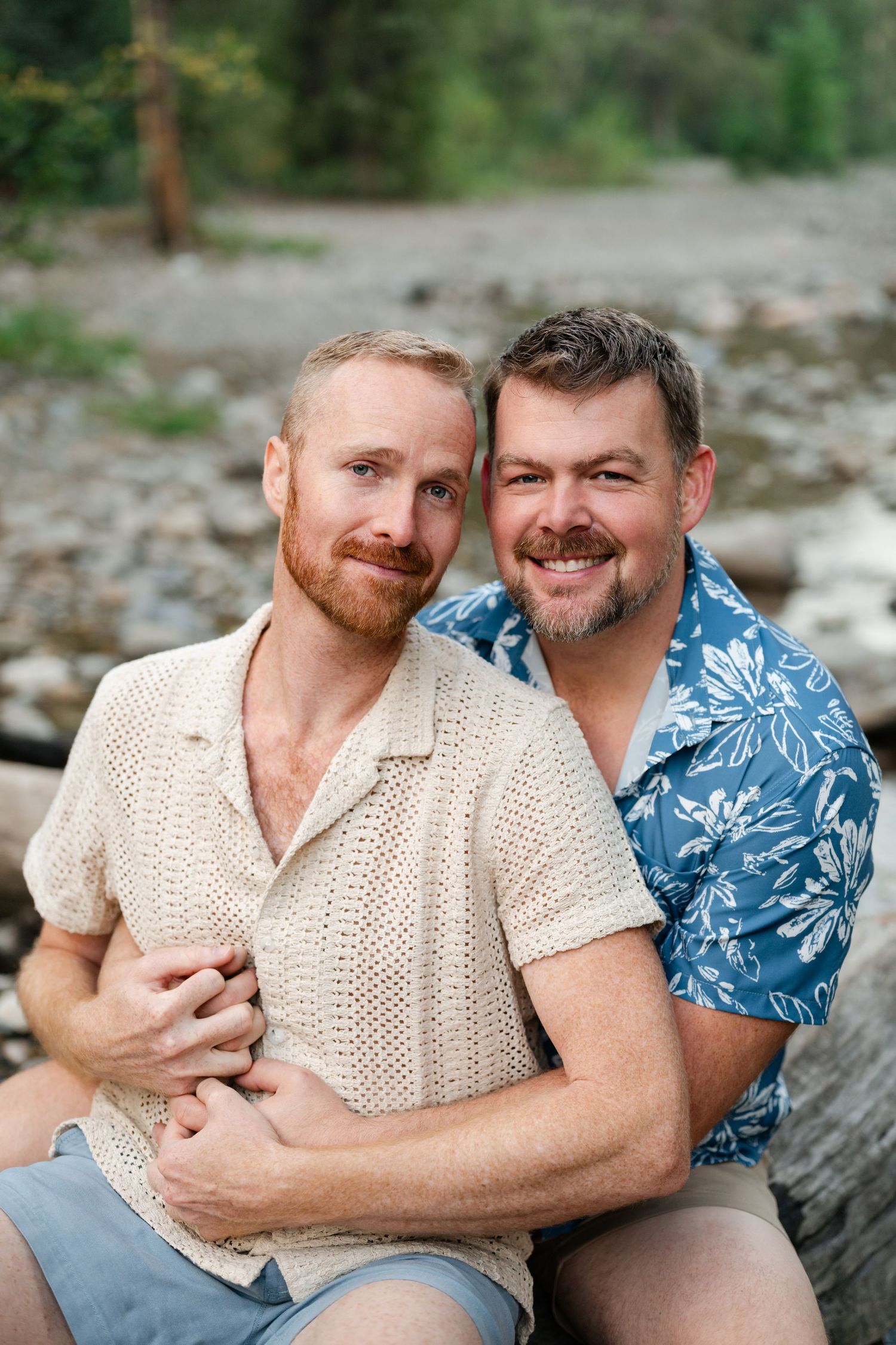 Two men embrace and smile outdoors near a rocky riverbed, LGBTQ friendly engagement photographer in Utah.