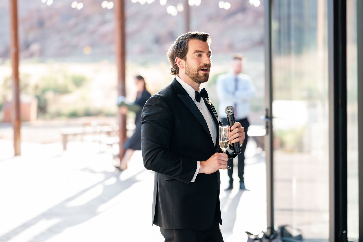Man in black tuxedo holding microphone giving speech at elegant indoor wedding venue.
