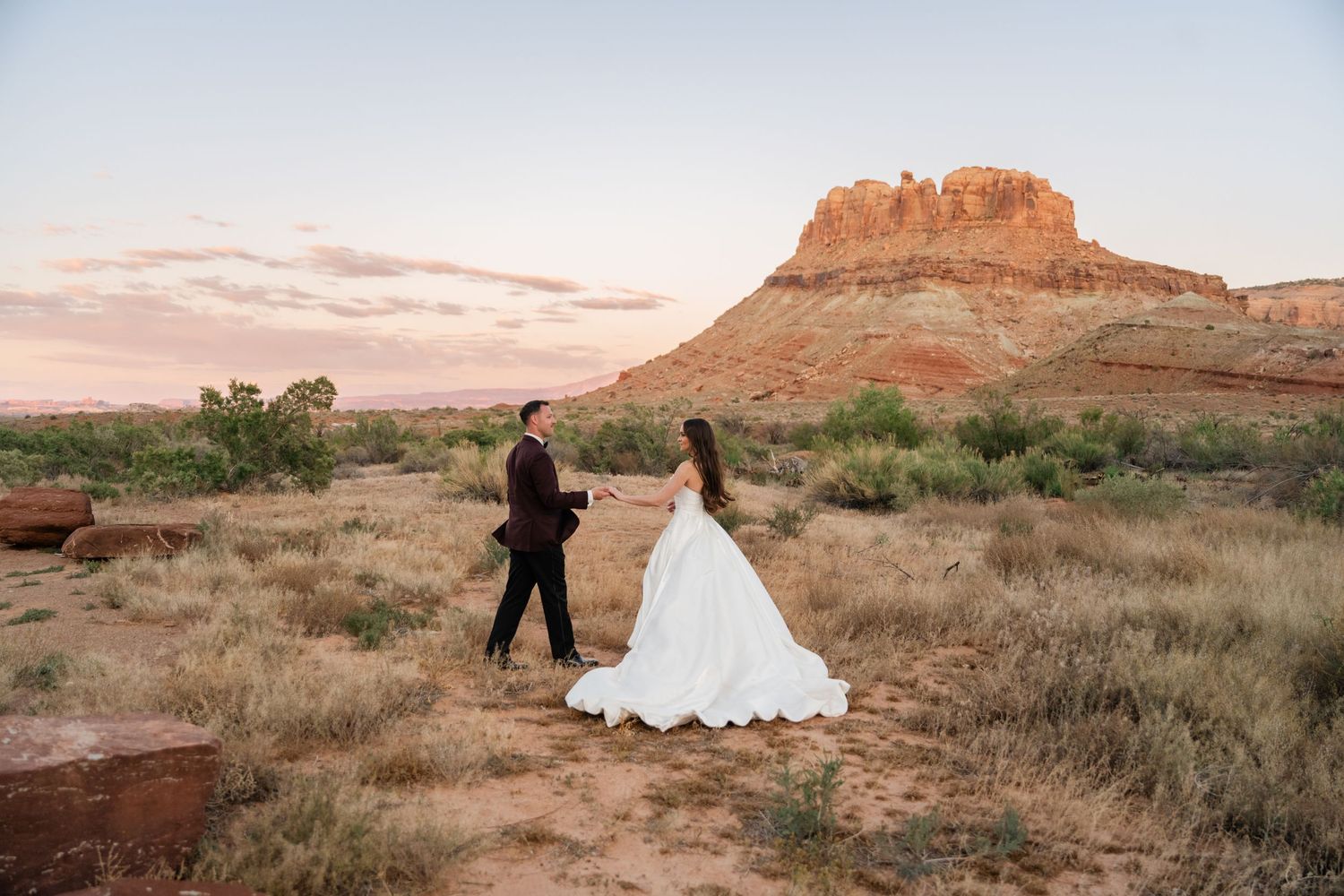 Bride and groom walk hand in hand through desert landscape with red rock butte at sunset.