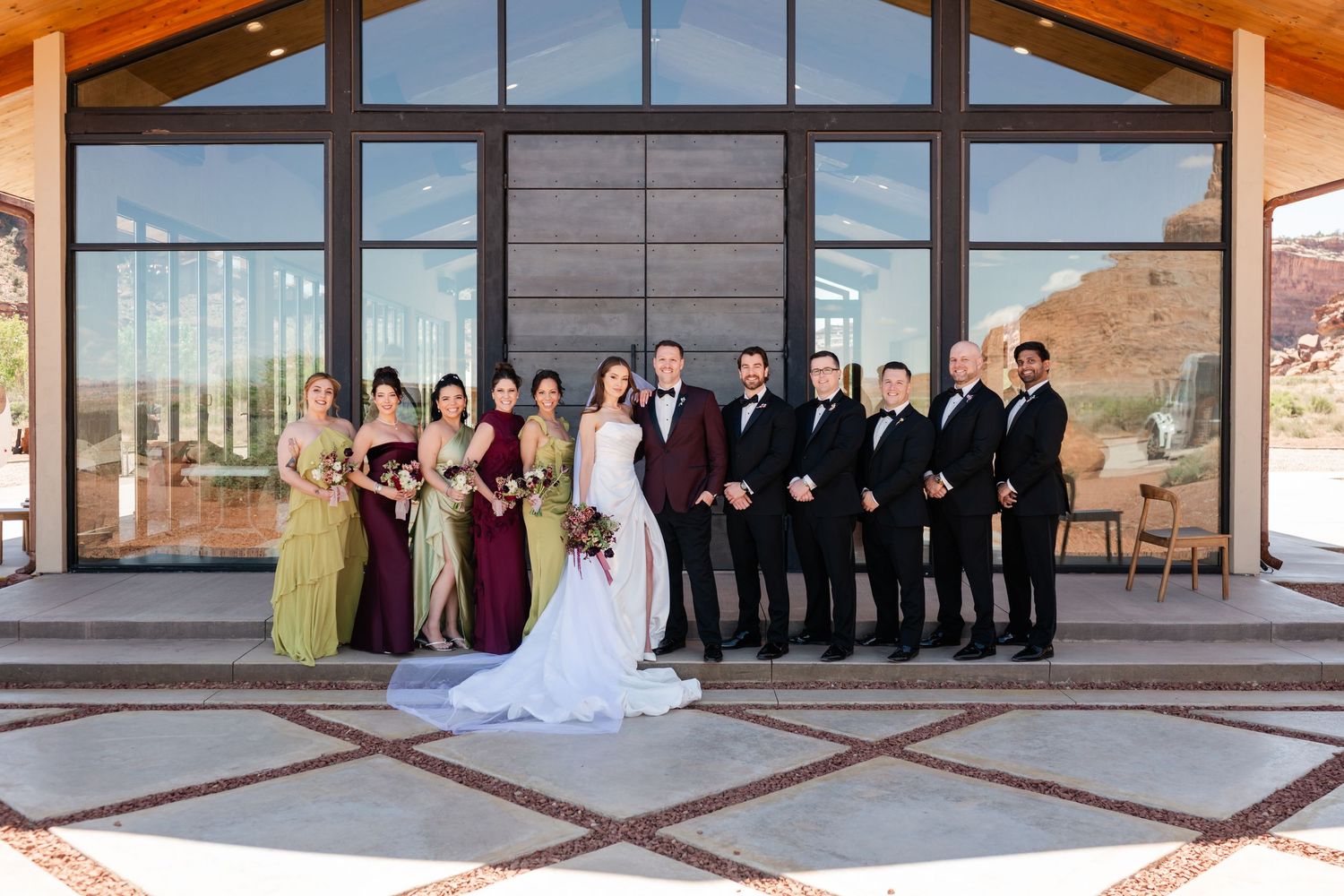 Wedding party poses outside The Red Earth Venue, bride in white gown with bridesmaids in jewel tones and groomsmen in black tuxedos.