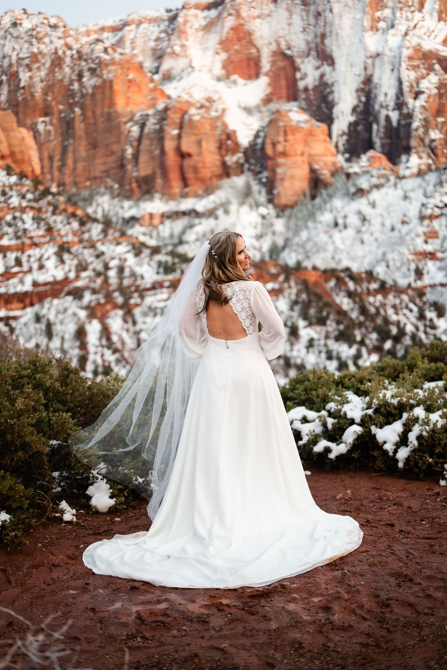 Bride in flowing white dress seated on red rocks with snowy cliffs in background.