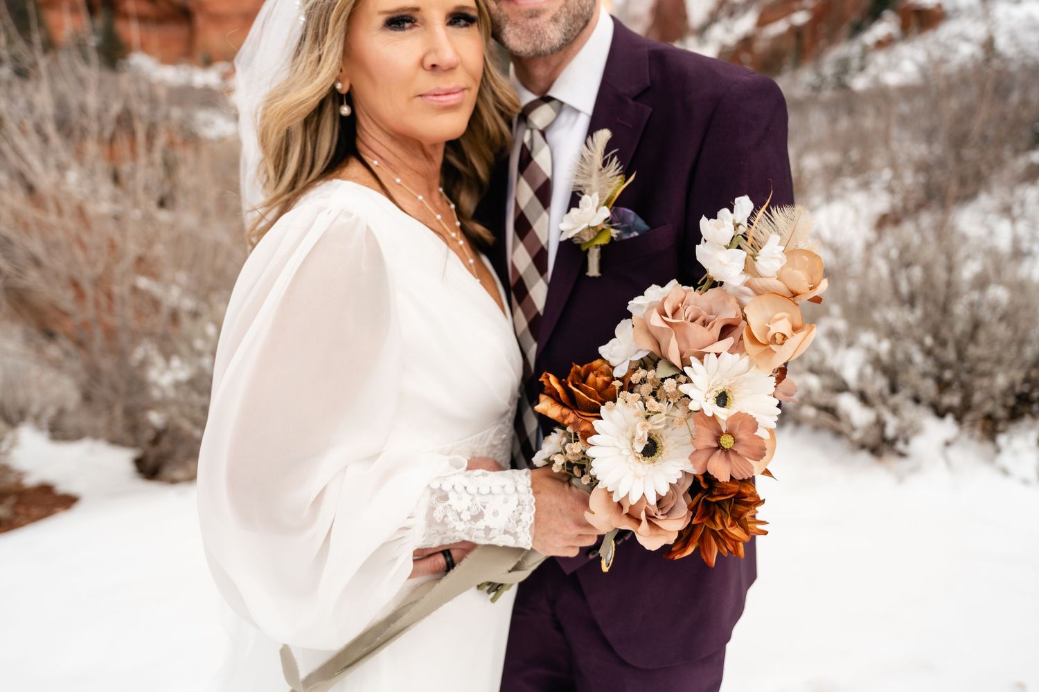 A romantic winter wedding photo shoot with a couple in formal attire posing in the snow with warm-toned floral bouquet.