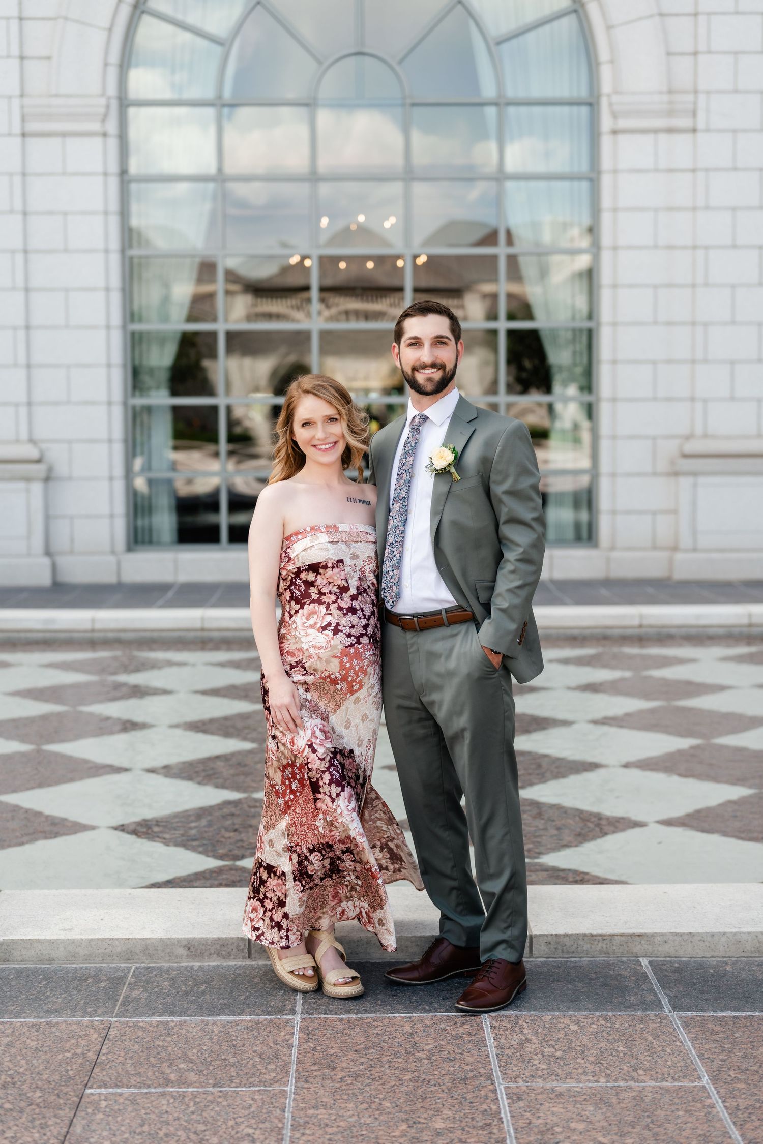 A couple poses together outside an elegant building with arched windows and checkered flooring.