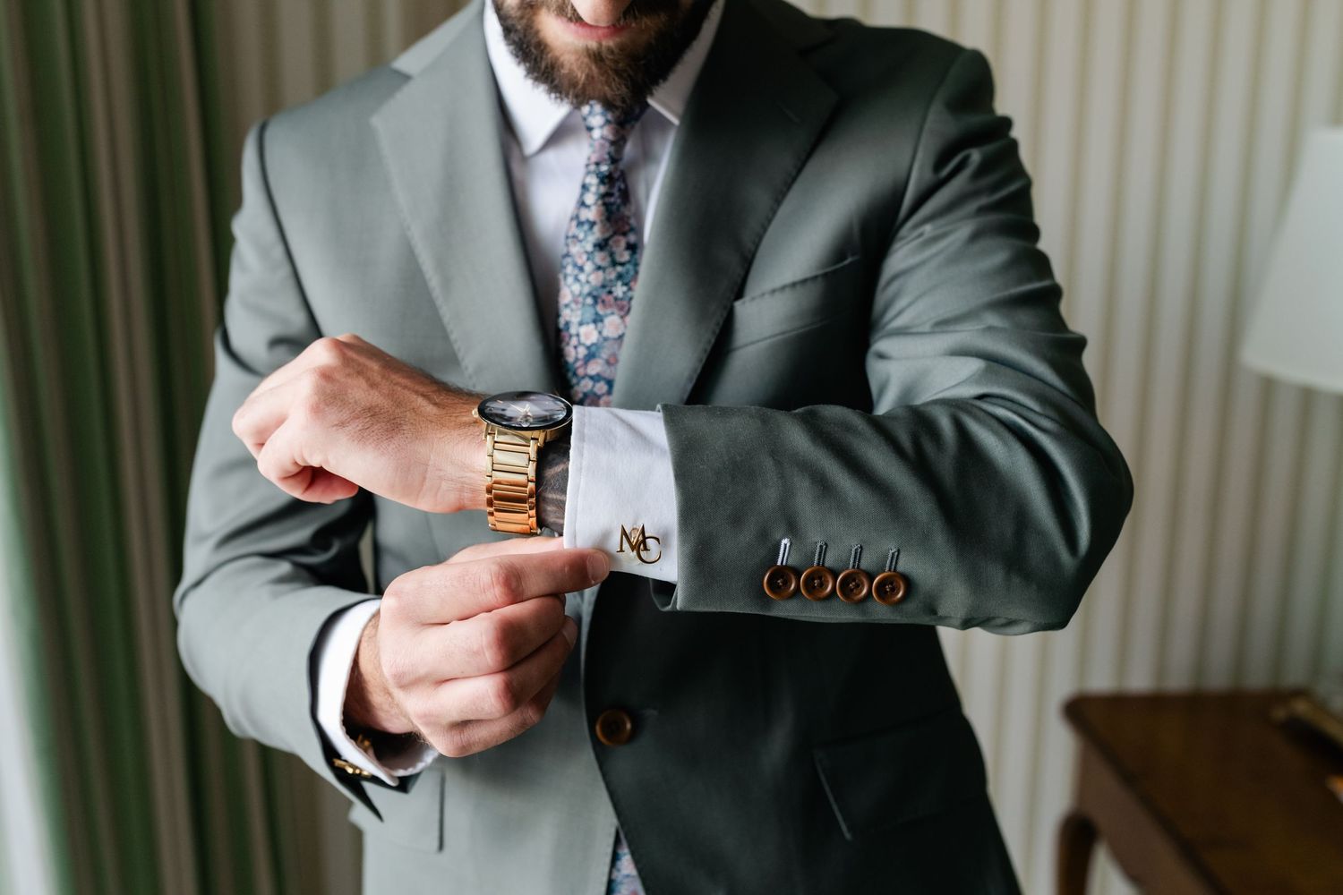 A person in a gray suit holds a glass of whiskey while getting ready for a formal event.