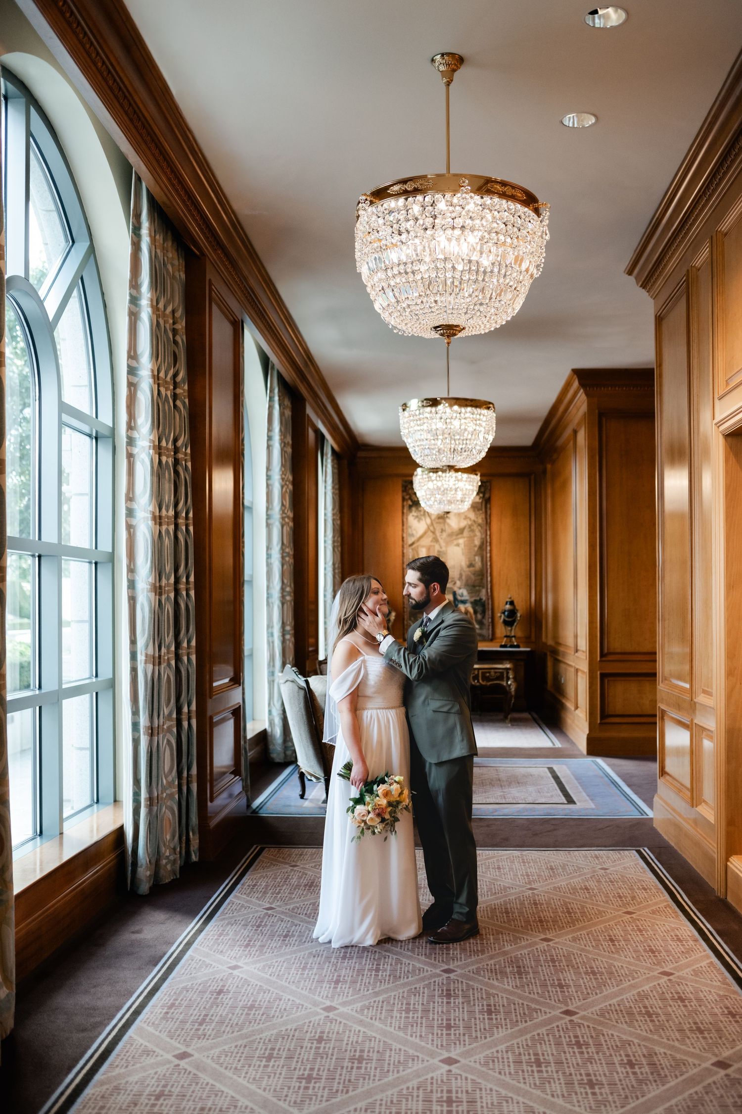 Wedding couple in formal attire stand in an opulent hallway with crystal chandeliers and wood-paneled walls.