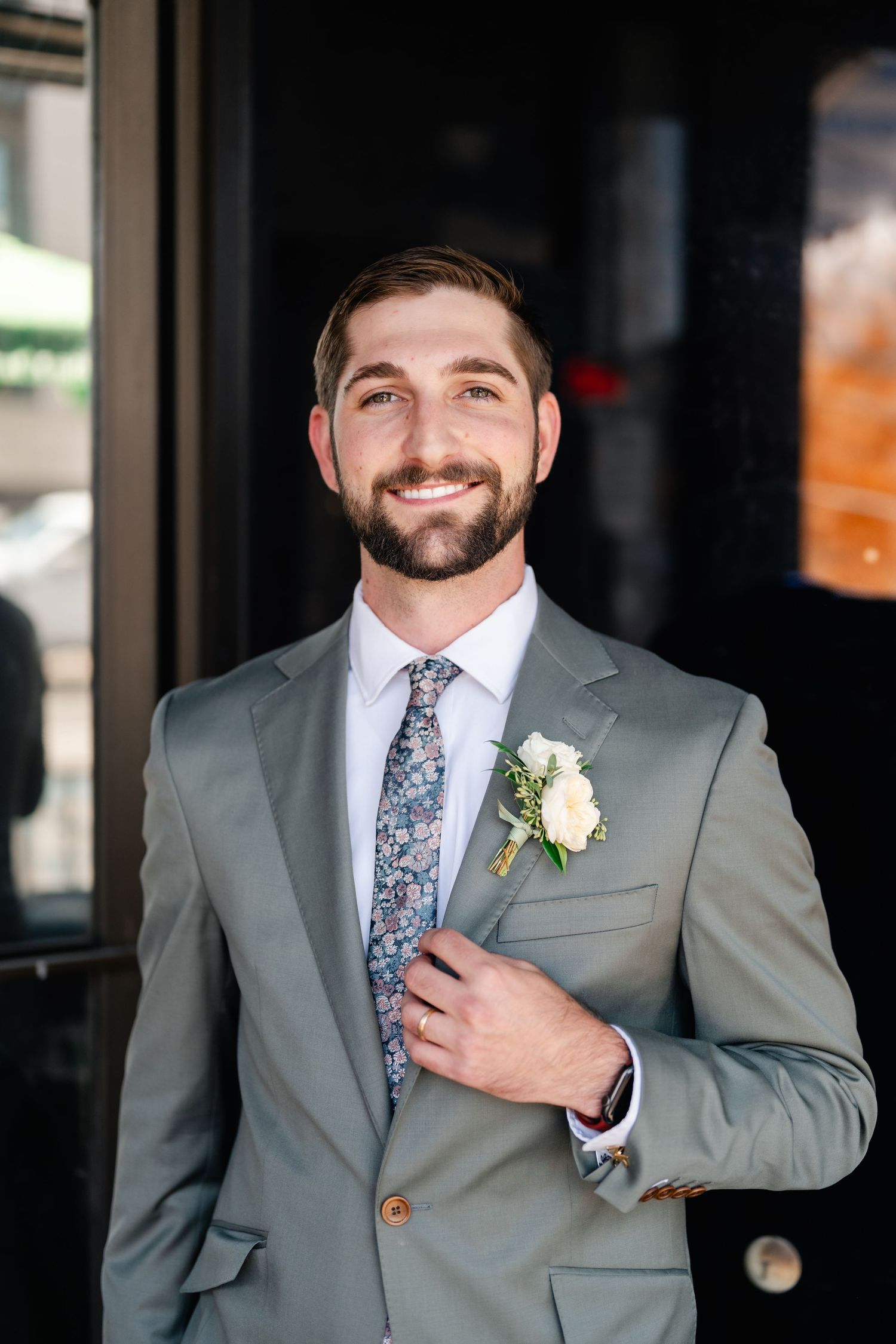 A person in a gray suit with a floral tie and boutonniere smiles while adjusting their jacket in a series of photos.