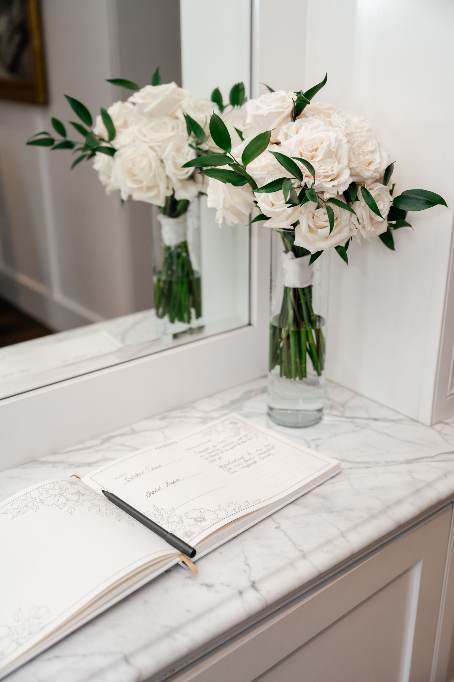 White roses in clear glass vases sit atop white marble surfaces in an elegant interior setting.