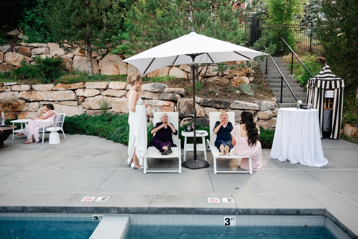 Outdoor wedding reception area with pool, white umbrellas, and stone retaining walls in a garden setting.