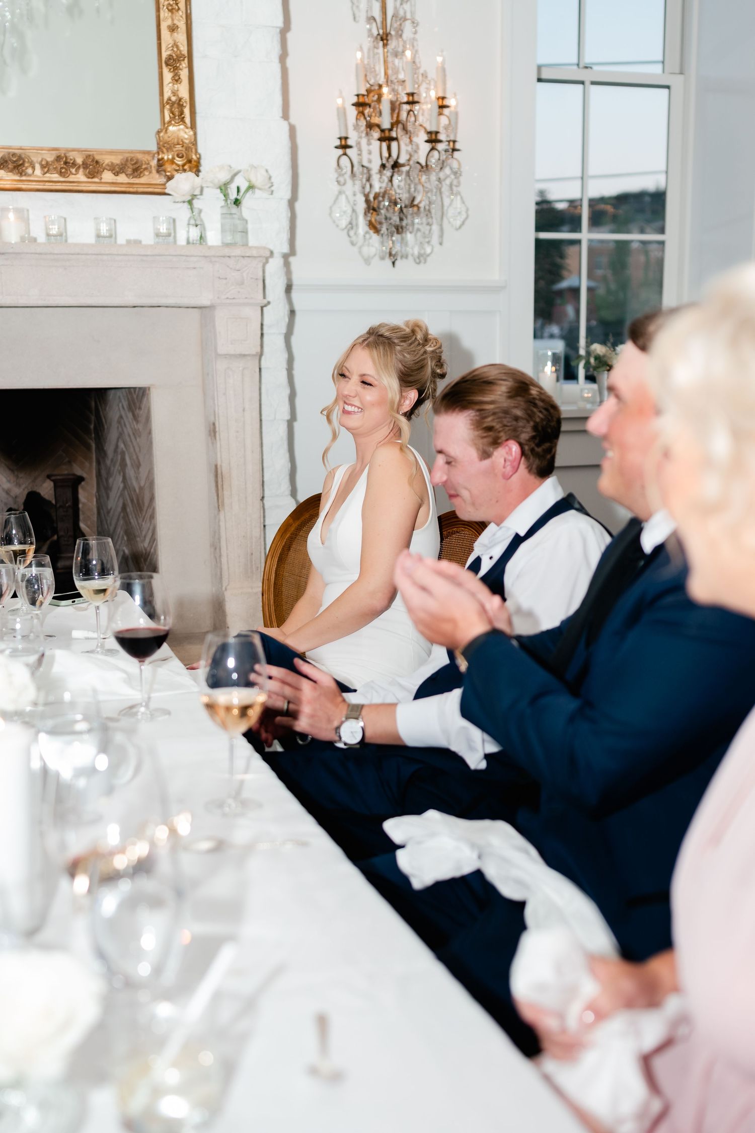Wedding guests enjoying reception dinner at elegant white tables with chandeliers overhead.
