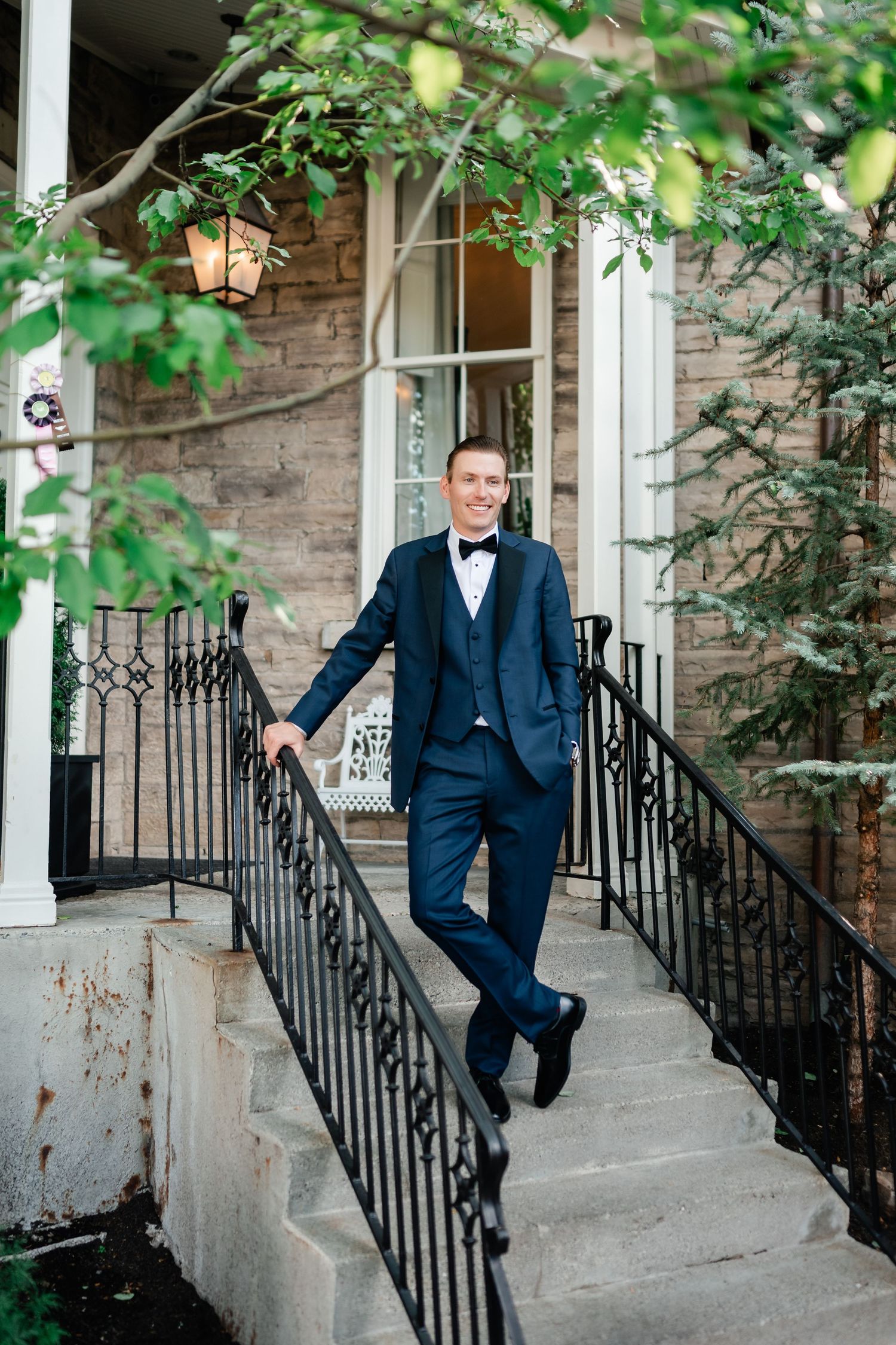 A groom in a navy suit poses on stone steps with black railings and green foliage in the background.
