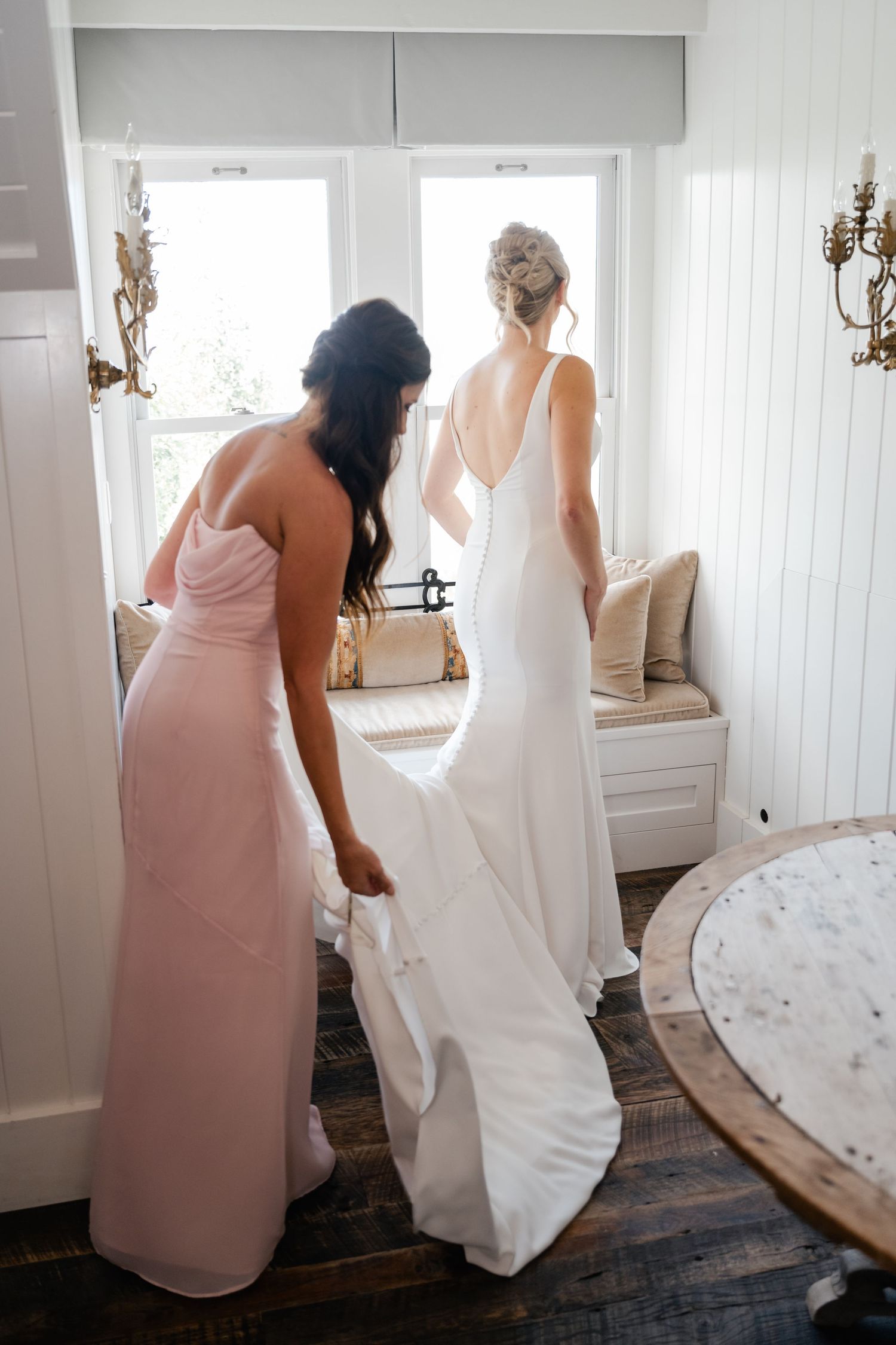 Wedding preparations showing flowing white dress and pink bridesmaid gown in a bright interior room.