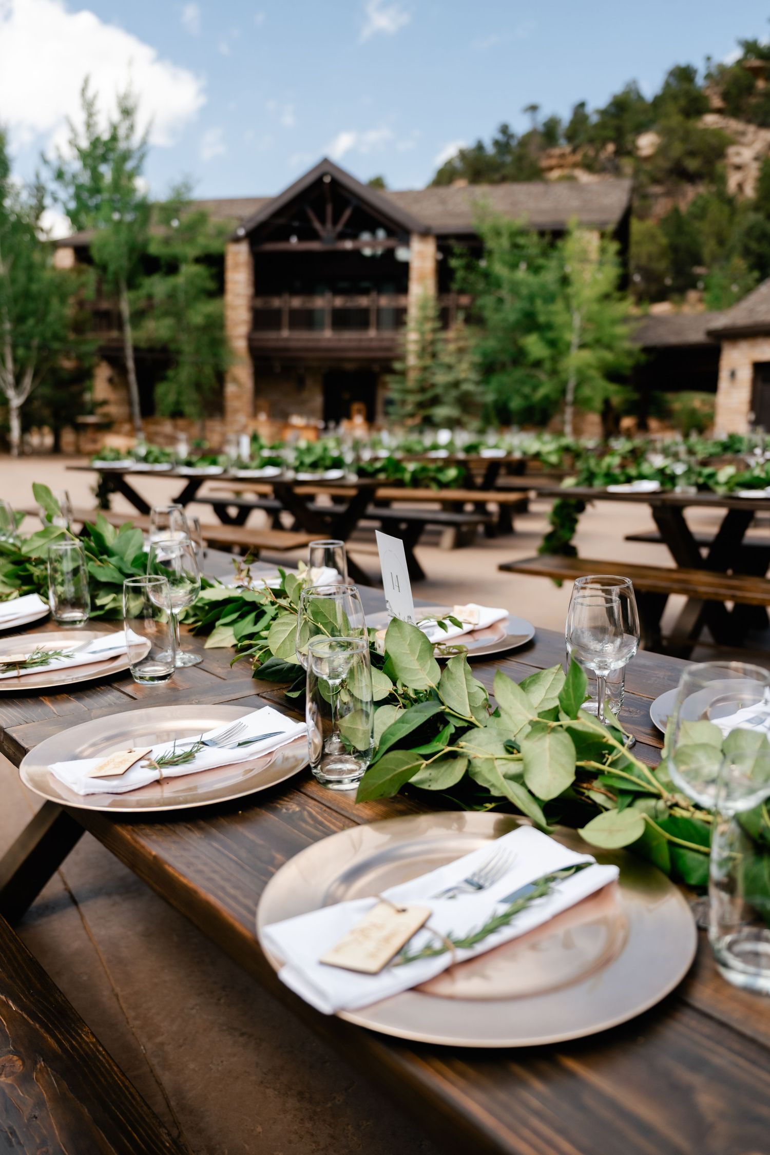 Elegant rustic wedding table settings with white plates and greenery garland on dark wooden tables at an outdoor venue.