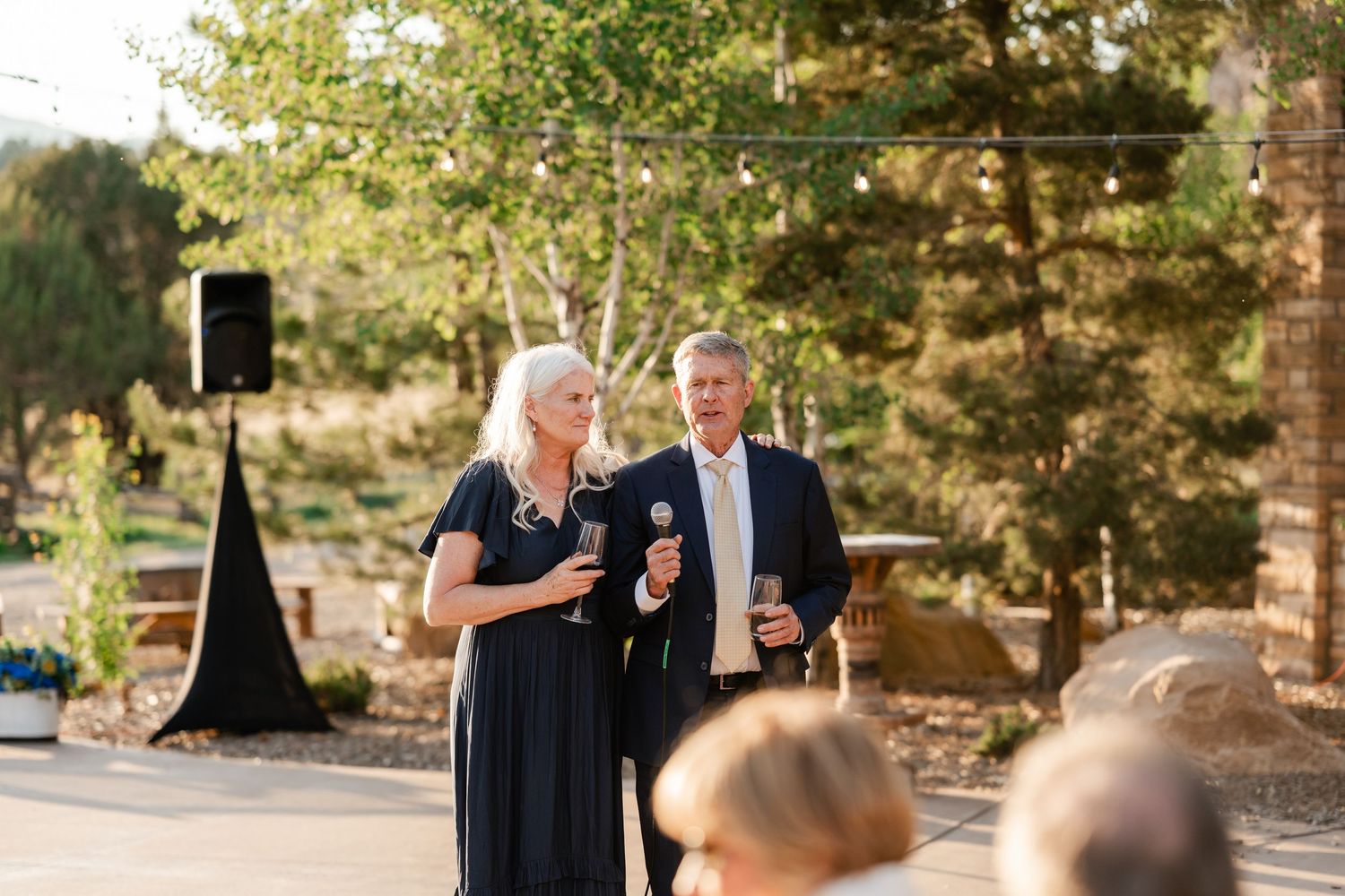 Speakers address wedding guests during outdoor ceremony with natural backdrop.