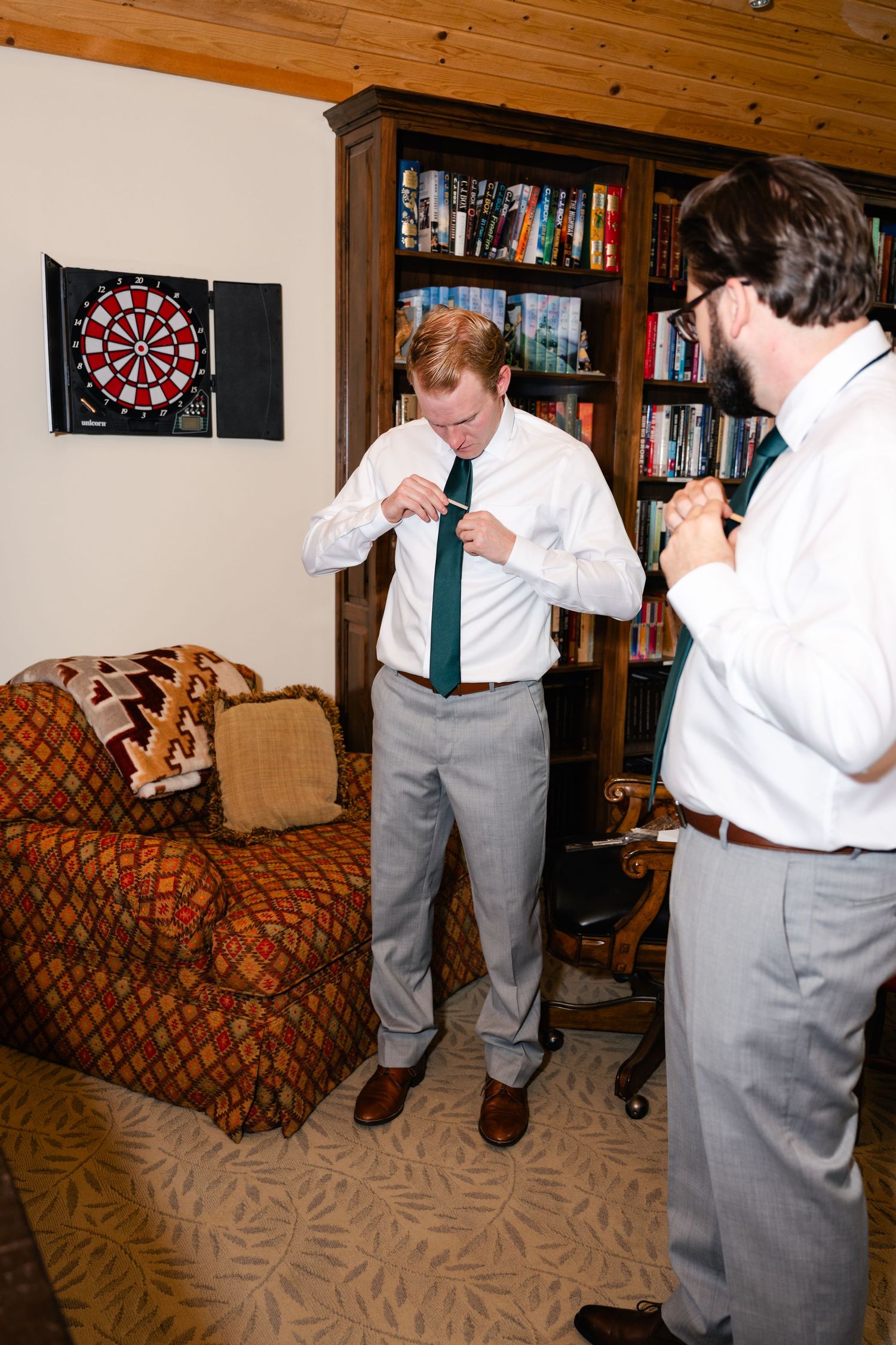 Two people in dress shirts and ties adjusting clothing near a dartboard and bookshelf.