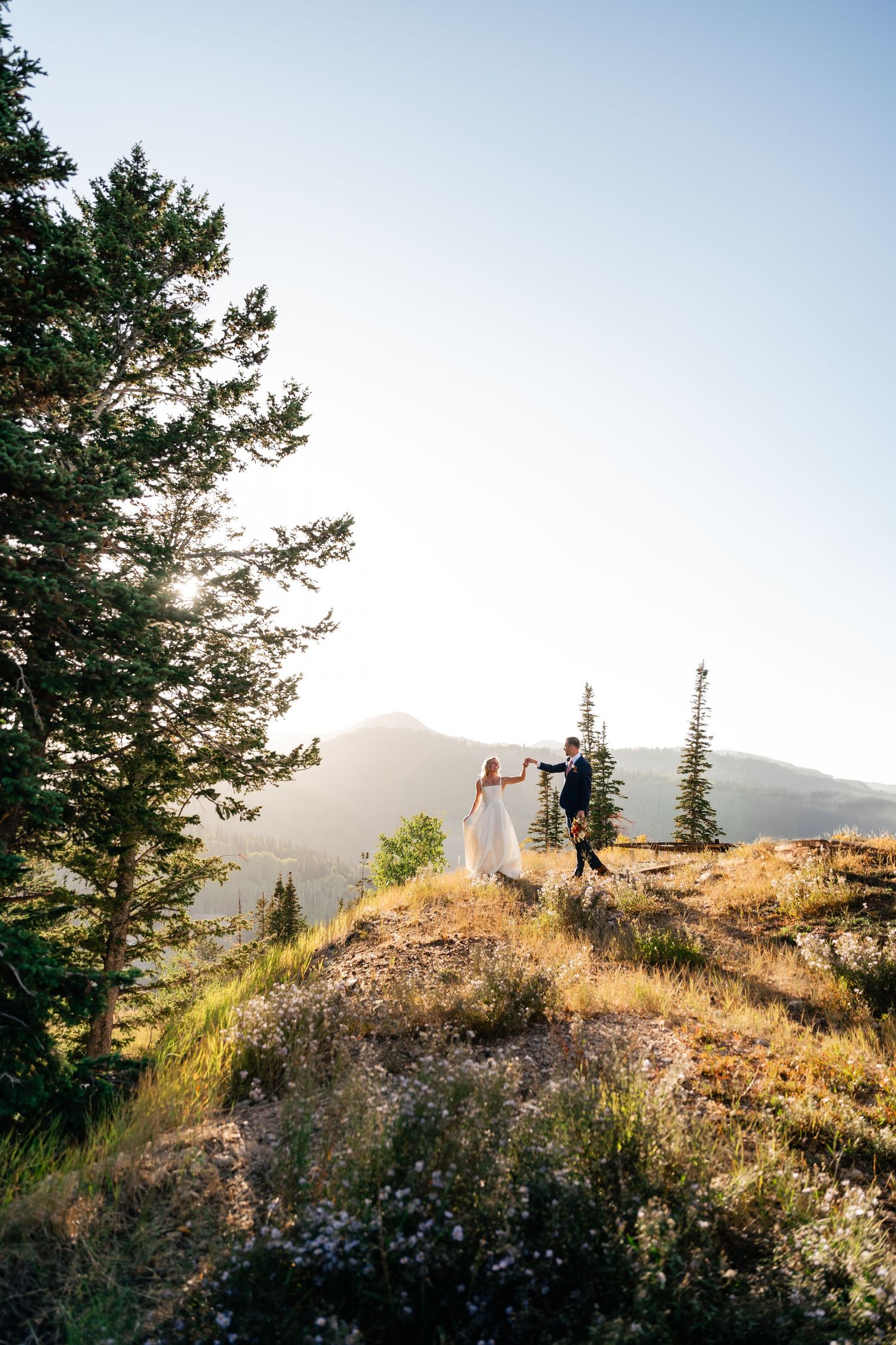 Silhouettes of people standing on a scenic mountain ridge at sunset with evergreen trees in the background.