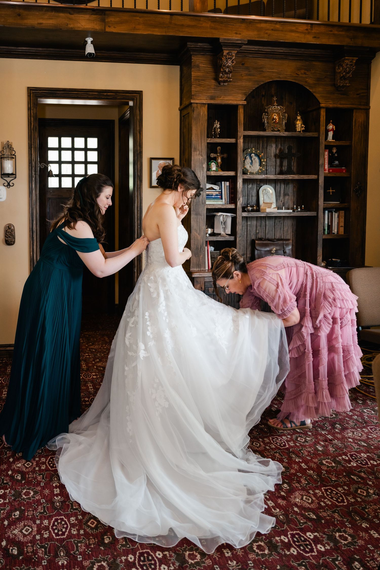 Wedding preparation scene showing flowing white bridal gown with attendants in navy and pink dresses.