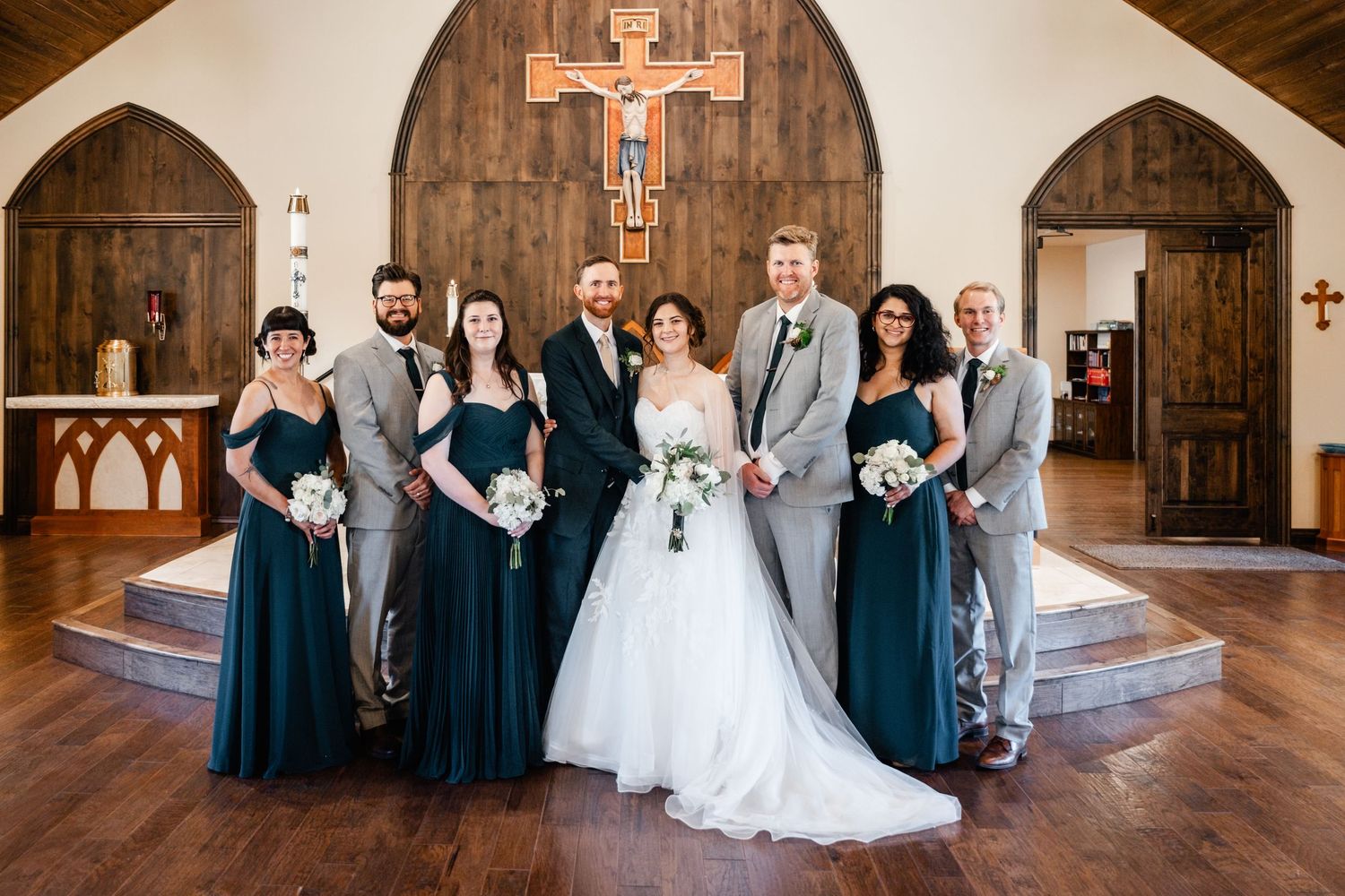 Wedding party in teal bridesmaid dresses and gray suits pose in front of wooden church altar with crucifix.