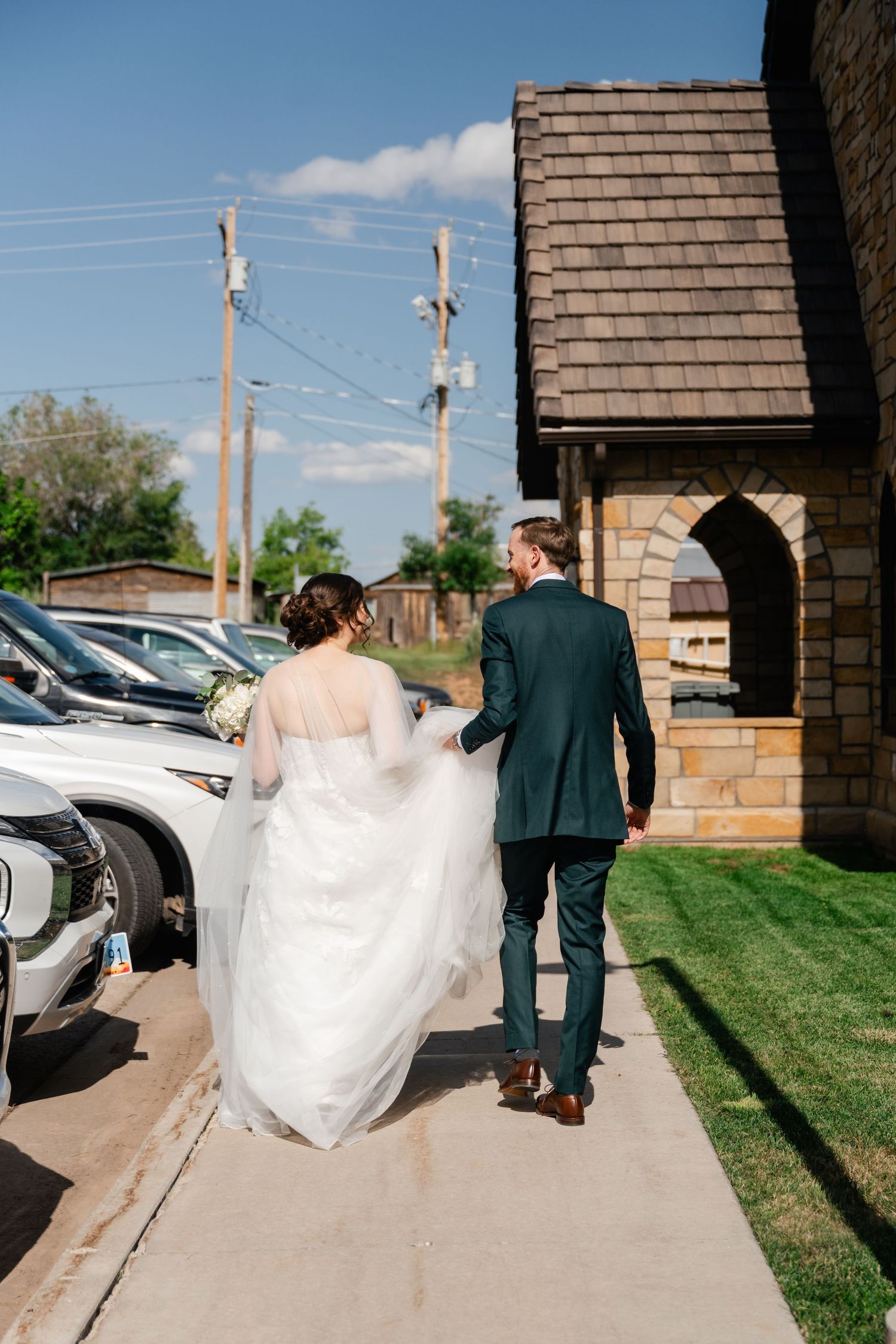 Wedding couple walks along a brick pathway outside with parked cars and buildings visible in the background.