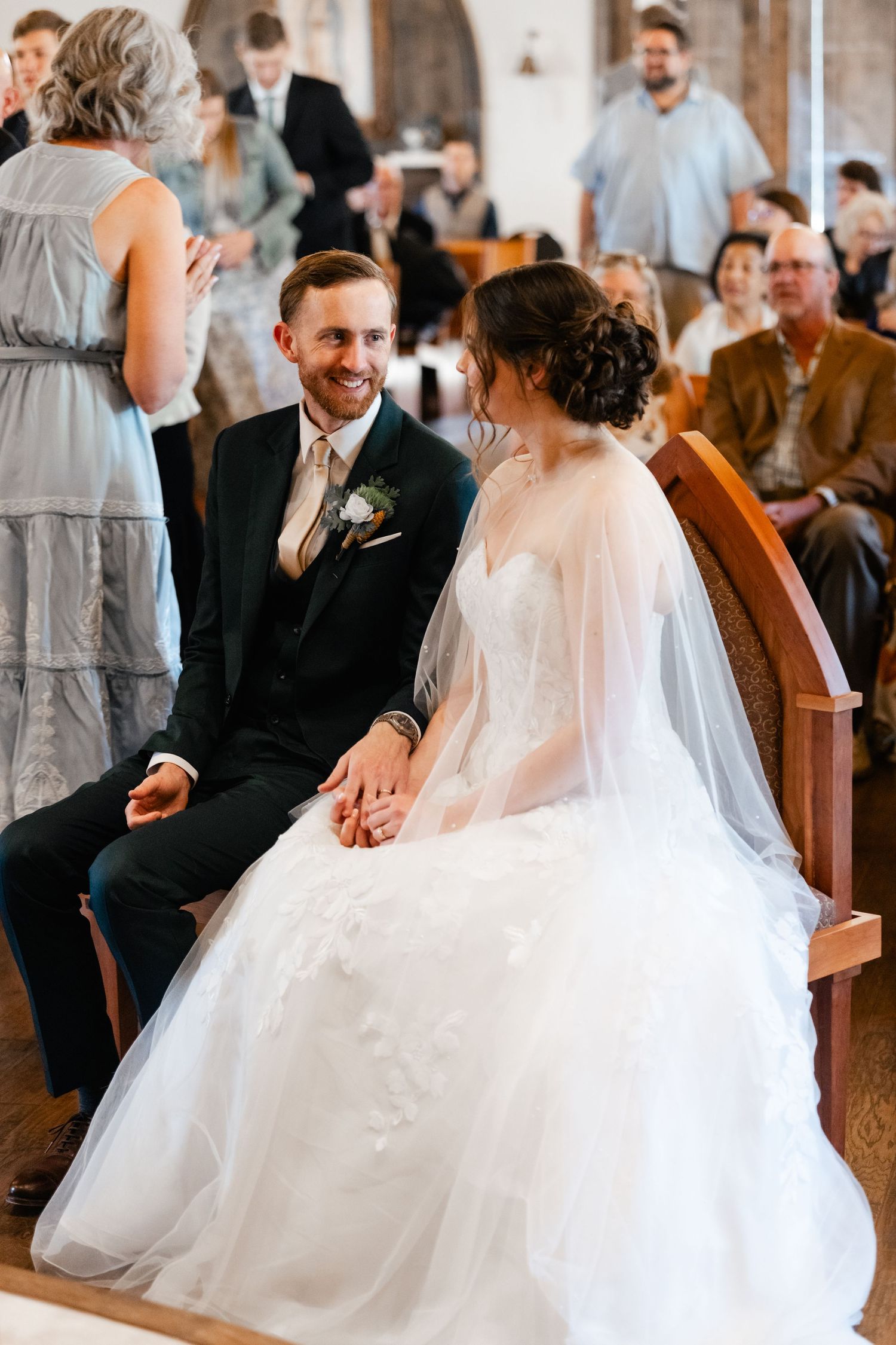 Wedding couple seated in wooden church pews during ceremony with guests in background.