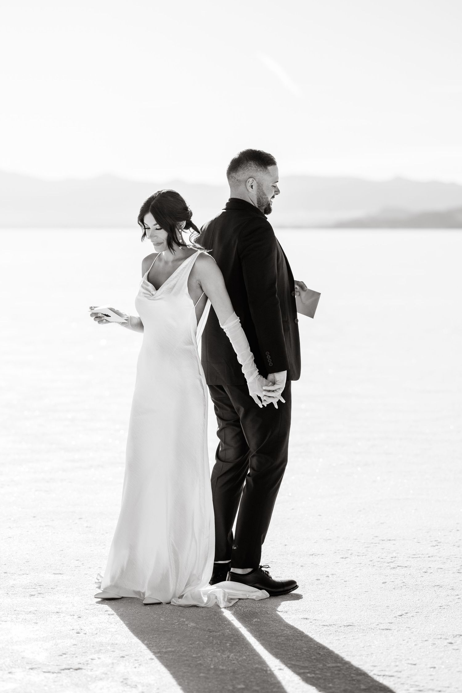 Black and white photo of a couple standing back to back on salt flats with dramatic shadows.