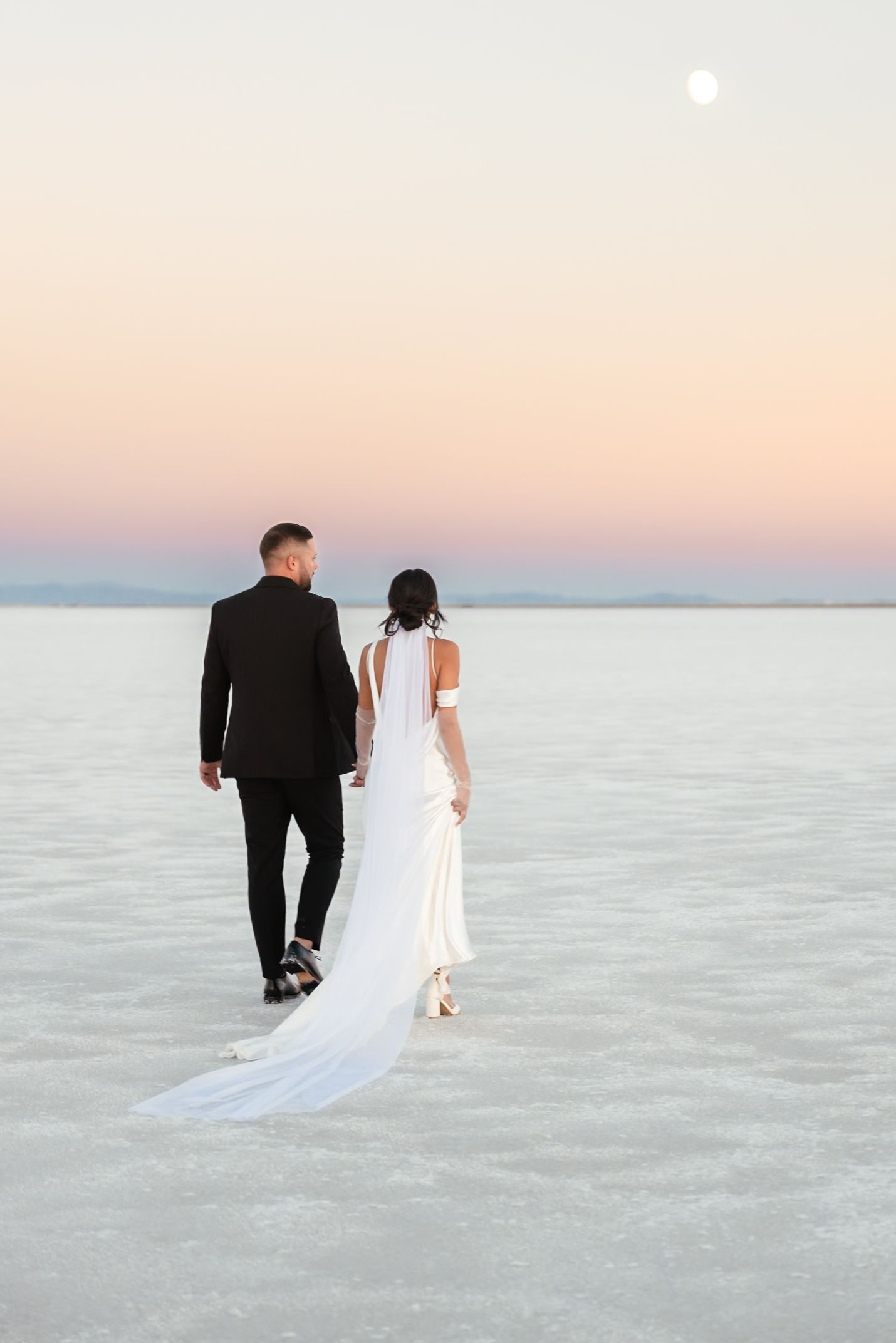 Bride and groom walking across the salt flats during sunset with trailing wedding dress.