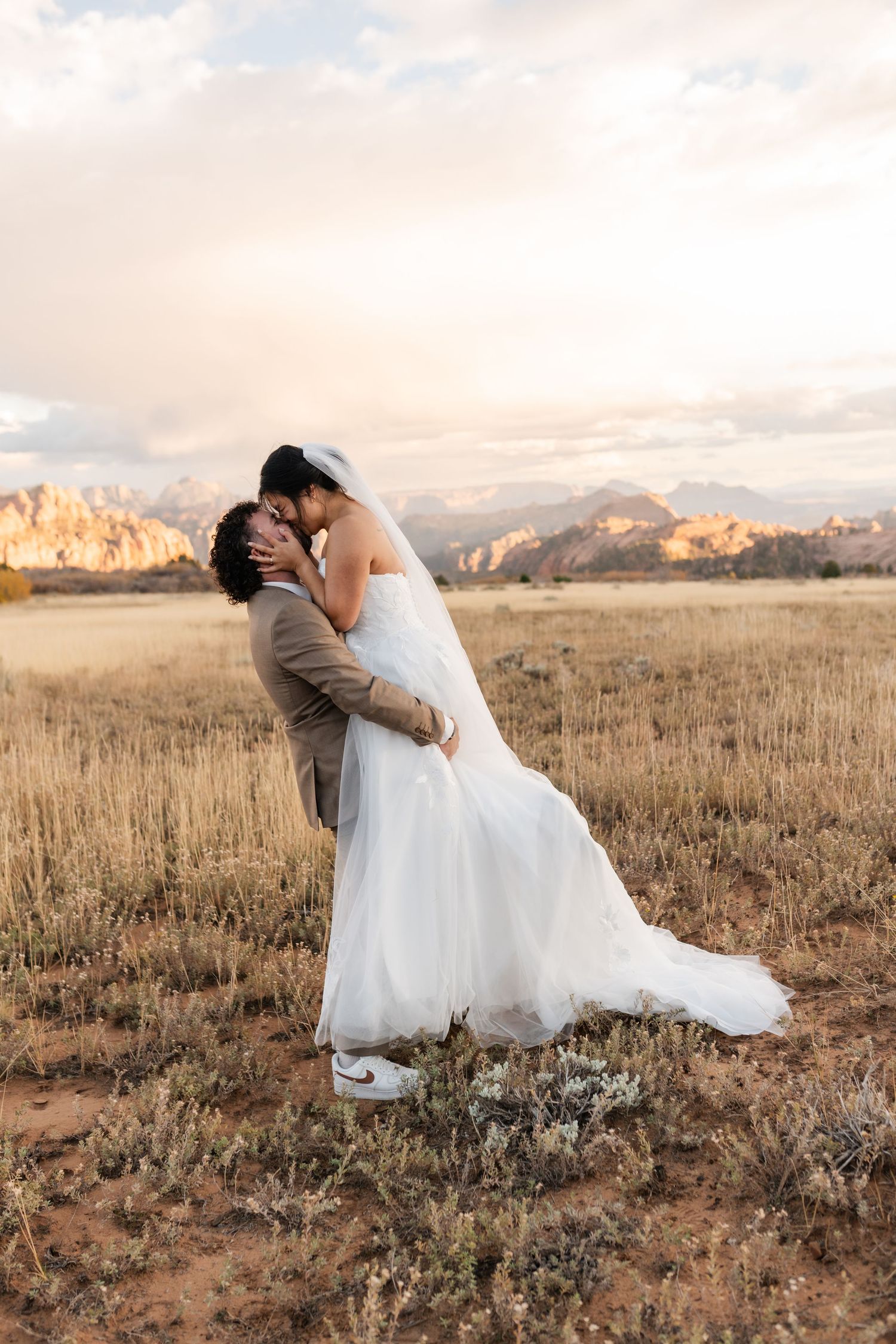 Newlyweds share an intimate moment in a desert field during sunset with red rock formations in the distance.