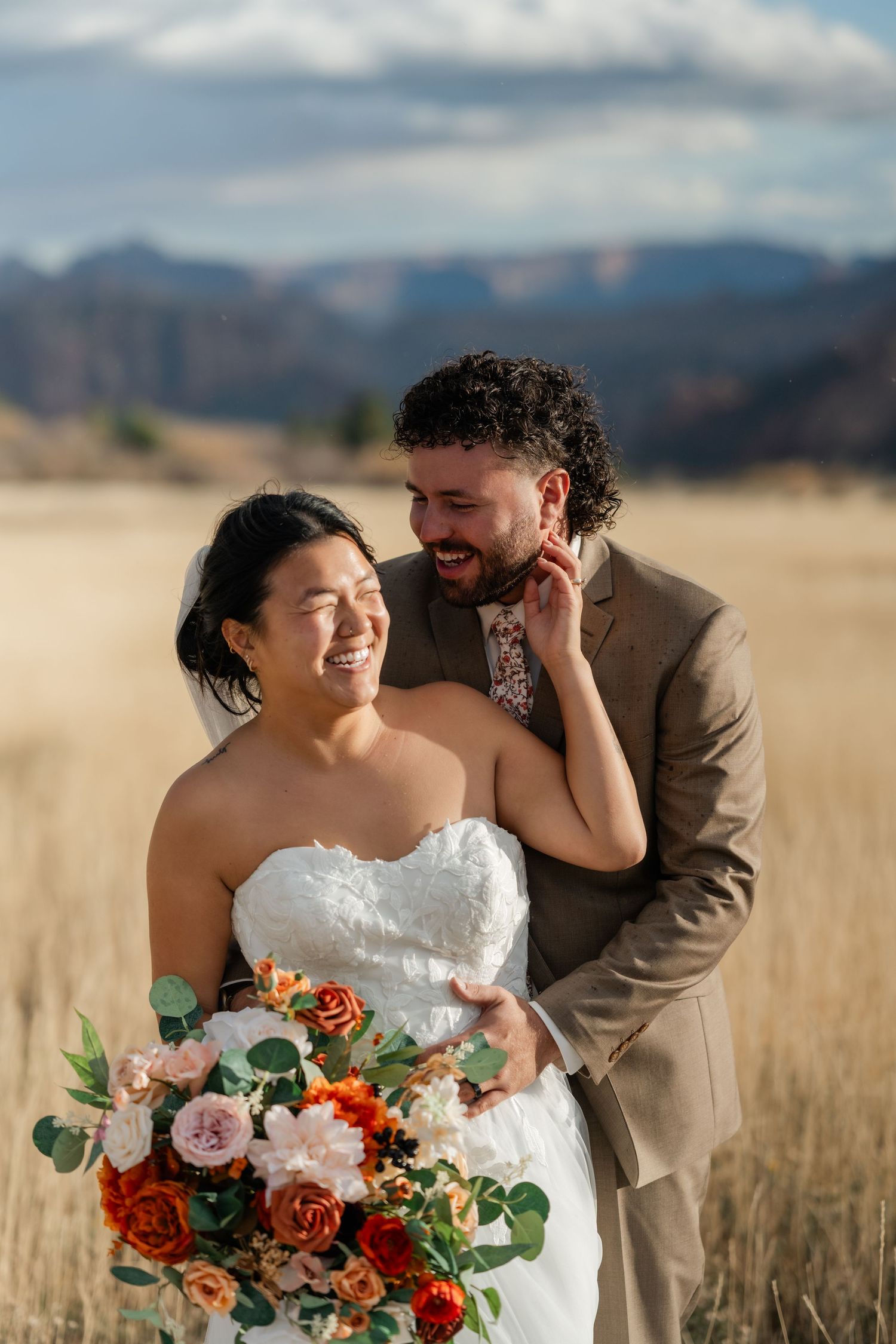 Newlyweds share an intimate moment in a rustic field with vibrant orange and pink bouquet.