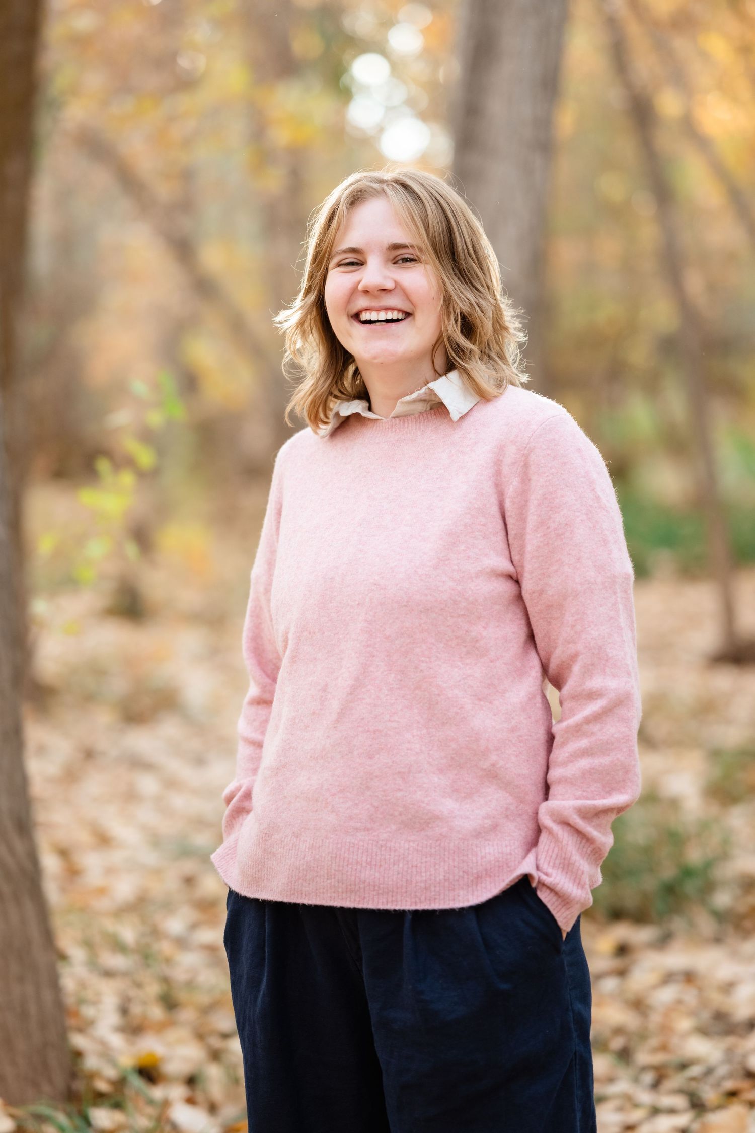 Person in pink and navy sweater poses in wooded area with fallen autumn leaves.