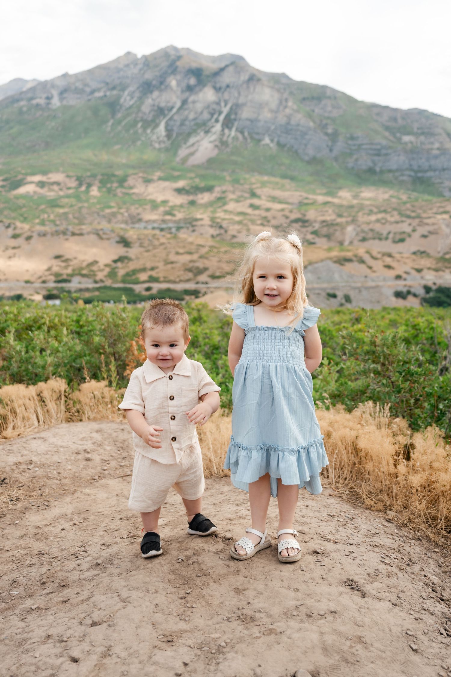 Two young children in light colored outfits standing on a trail with mountains in the background.