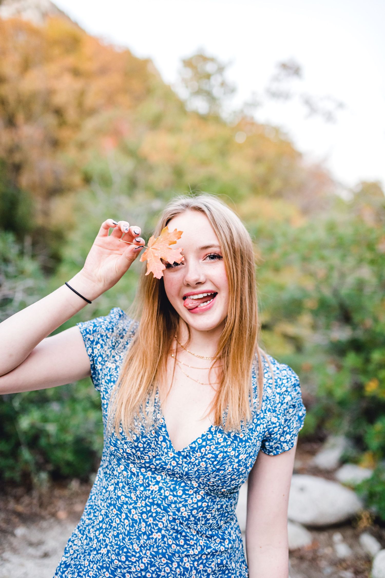 A person in a blue floral dress poses outdoors on a sunny day with autumn foliage in the background.