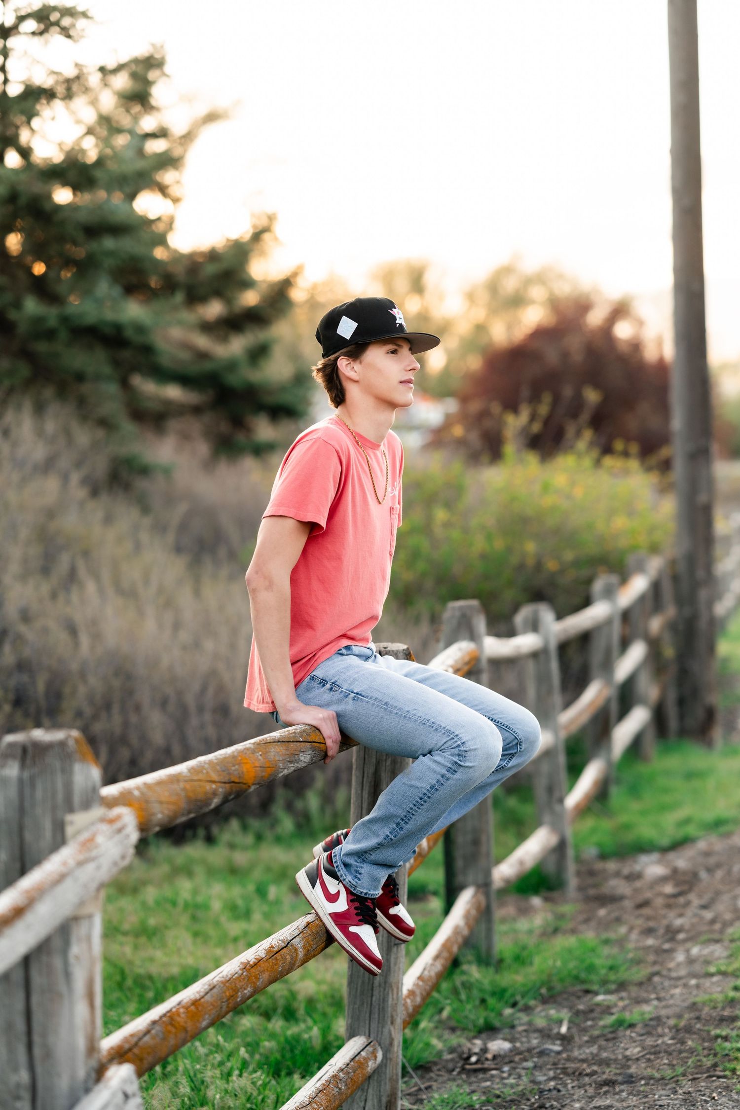 Person in pink t-shirt and jeans sits casually on wooden fence at sunset in rural setting.